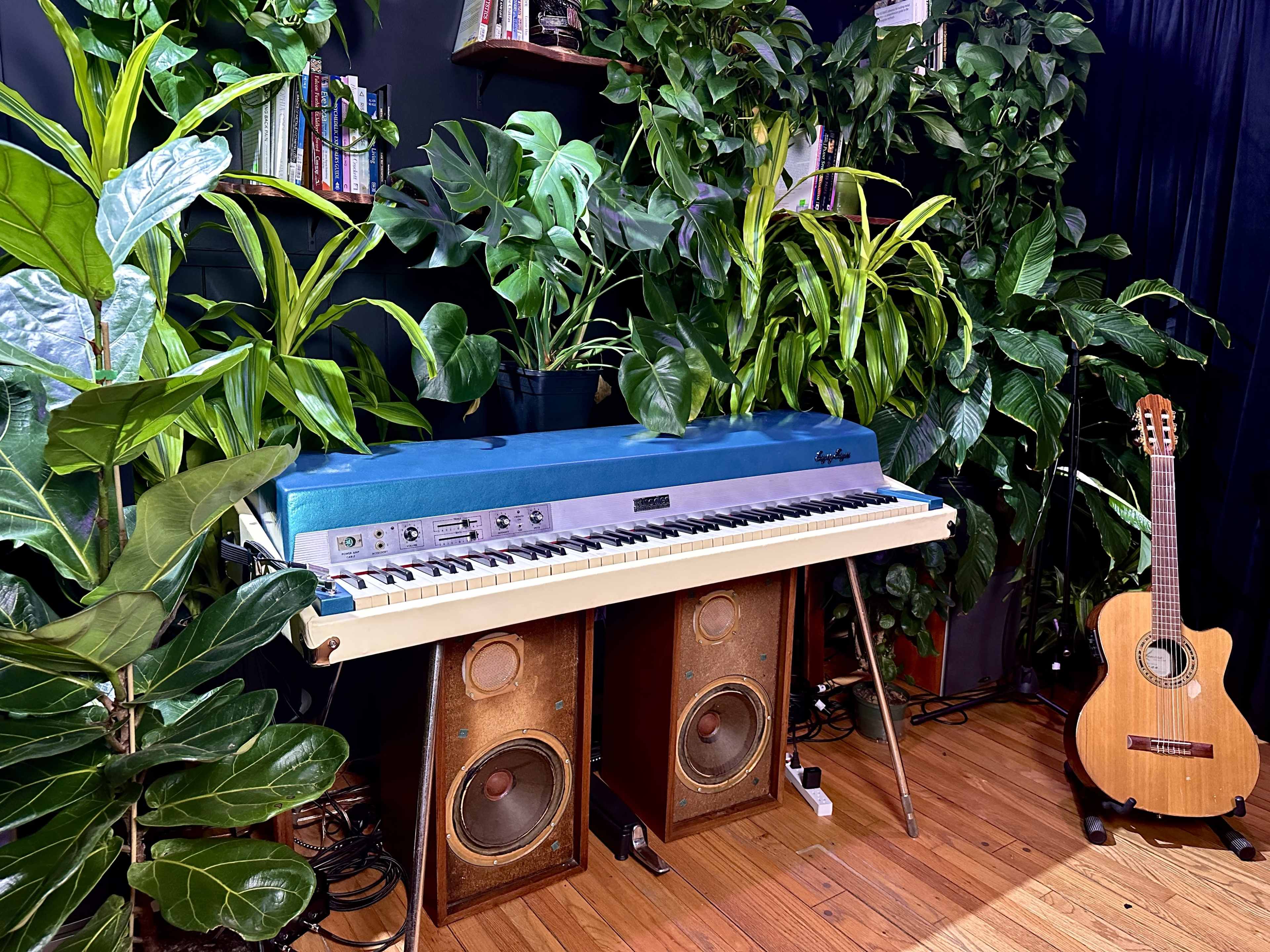 A blue electric piano is positioned between tall green plants on a wooden floor, with a guitar and vintage speakers nearby.