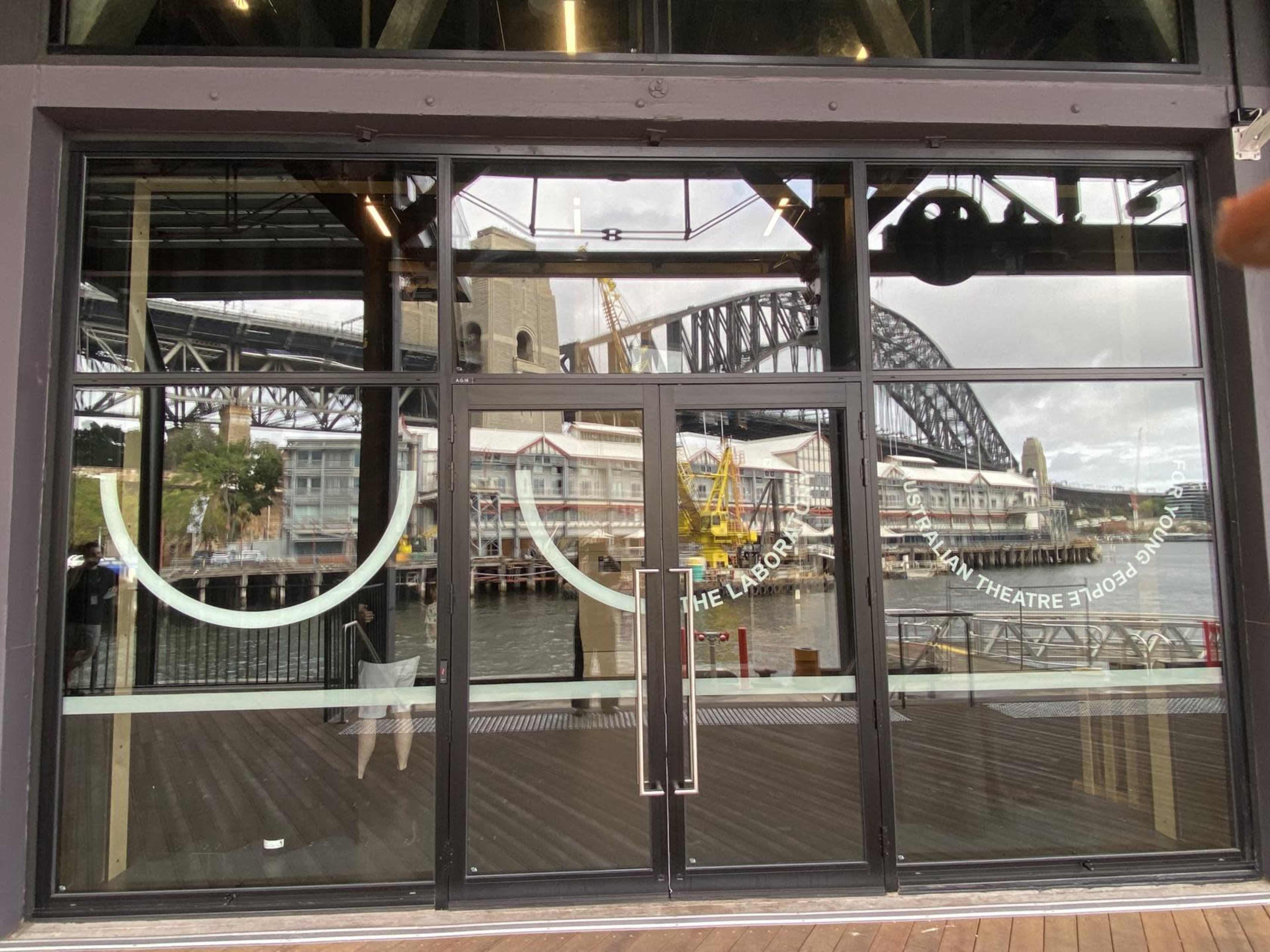The image shows a glass storefront with a view of the Sydney Harbour Bridge and nearby buildings reflected in the windows.