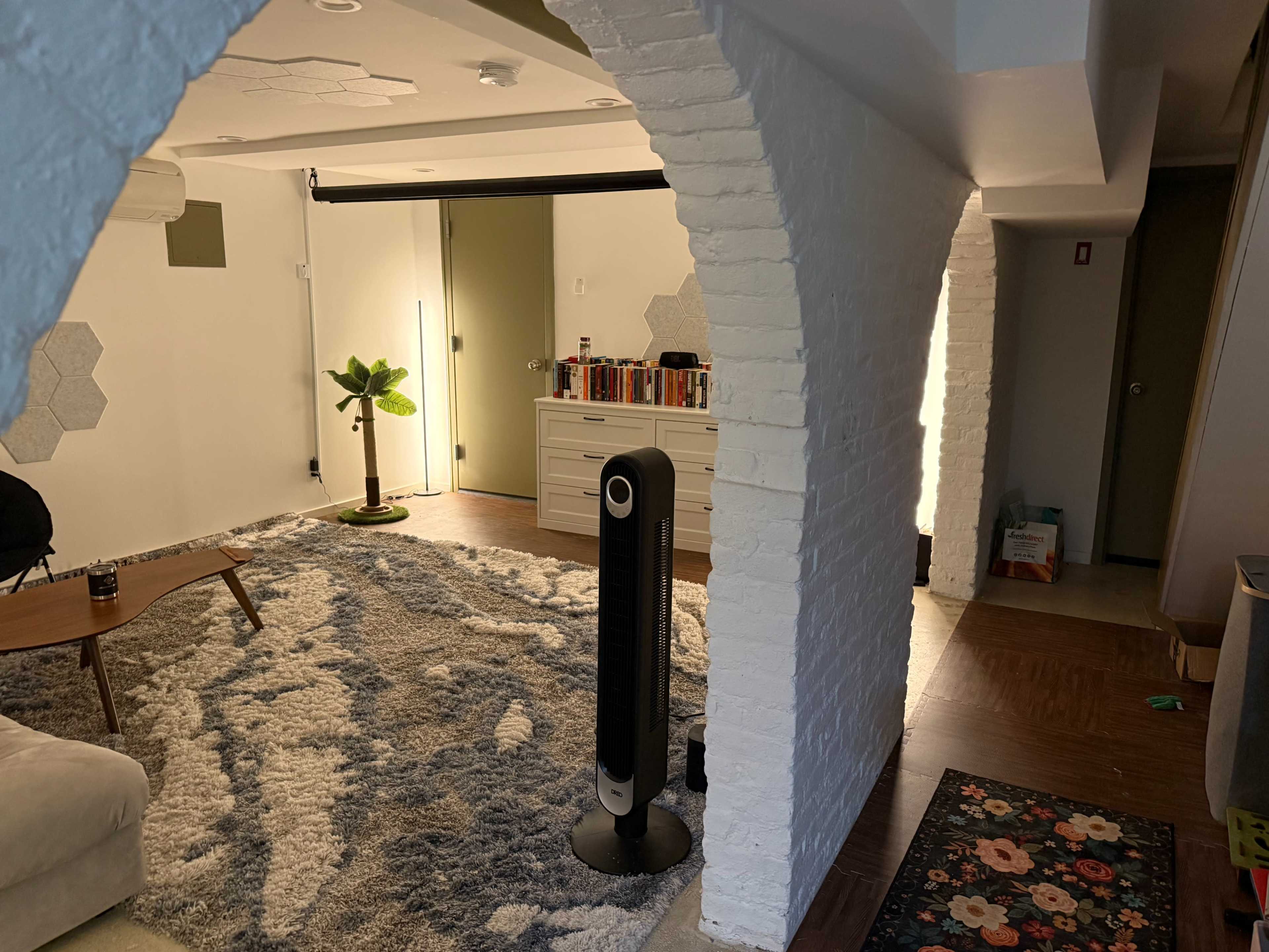 The image shows a modern interior space with an archway, featuring a cozy area with a large rug, a coffee table, a standing fan, and a bookshelf against the wall.