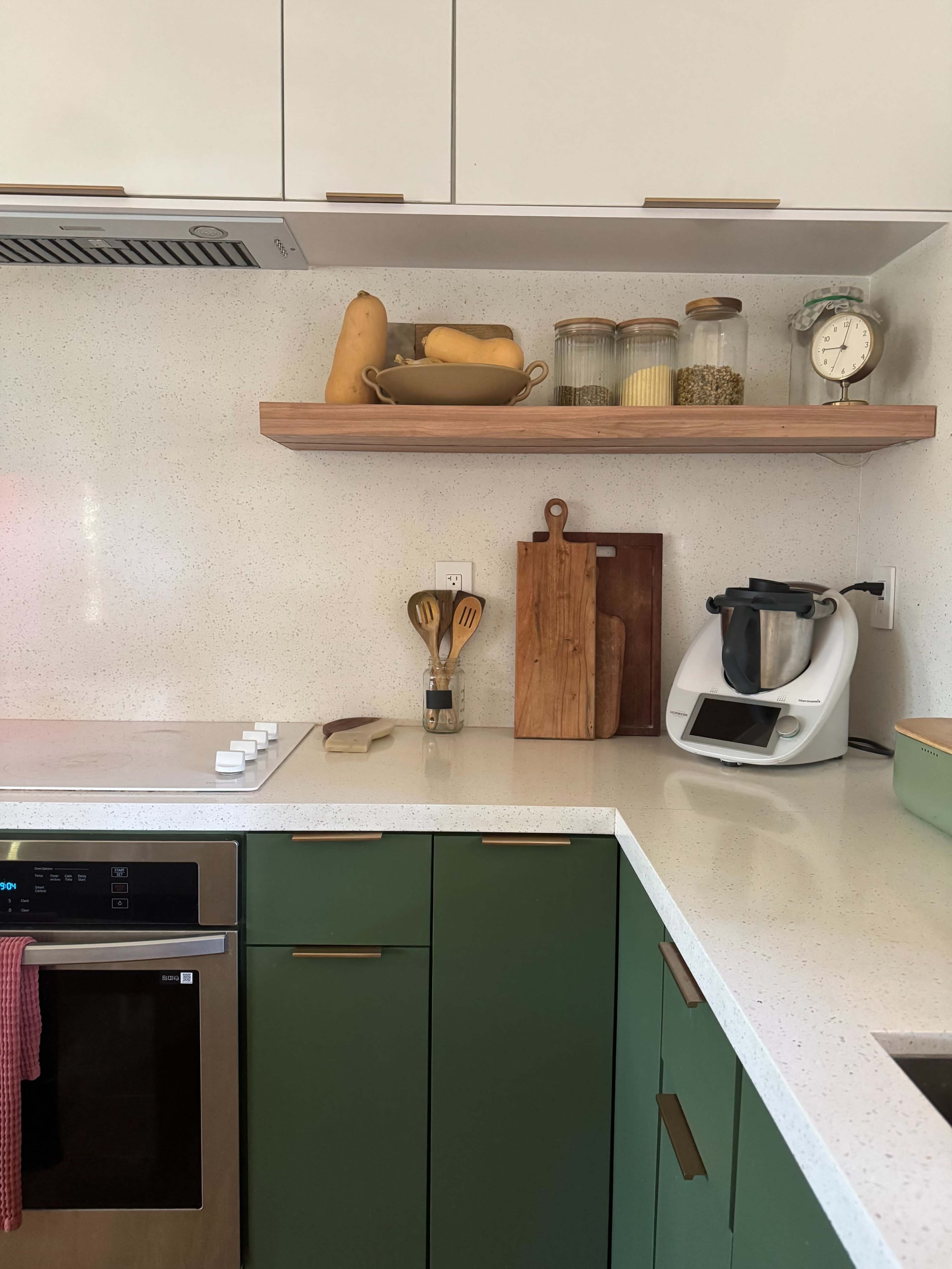 The image shows a modern kitchen with green cabinetry, a white countertop, stainless steel appliances, and open shelving displaying jars and utensils.