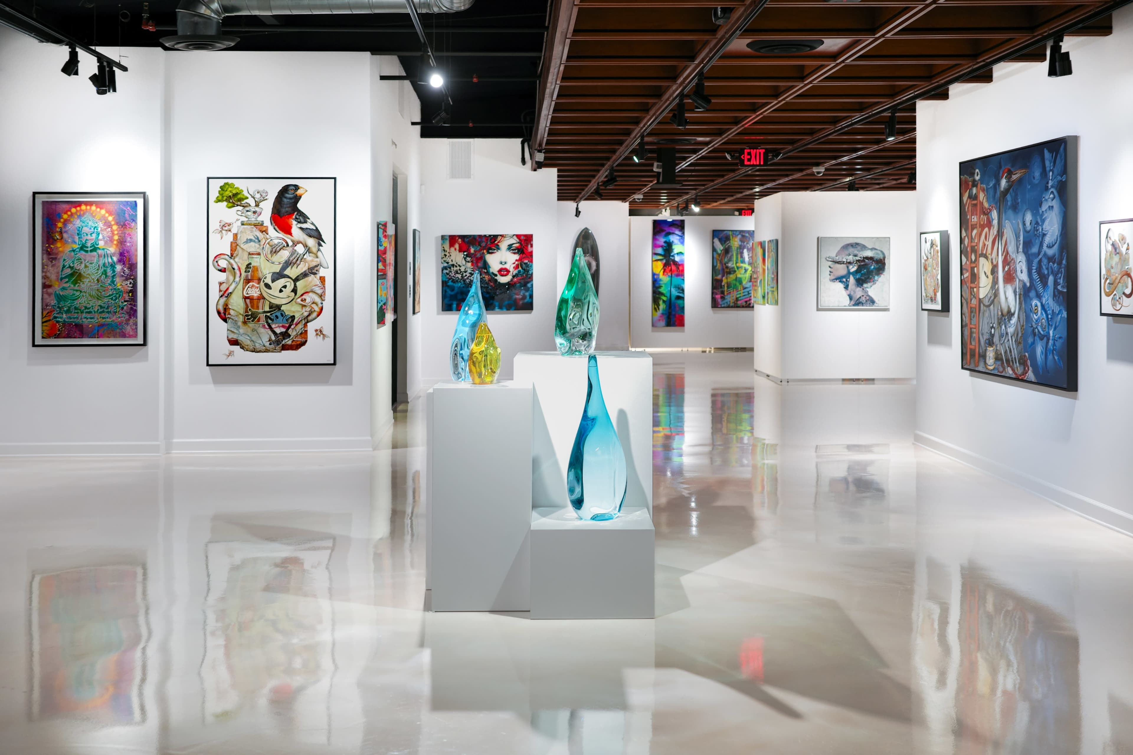 The image shows an art gallery featuring colorful paintings on walls and sculptural glass pieces displayed on a white pedestal.