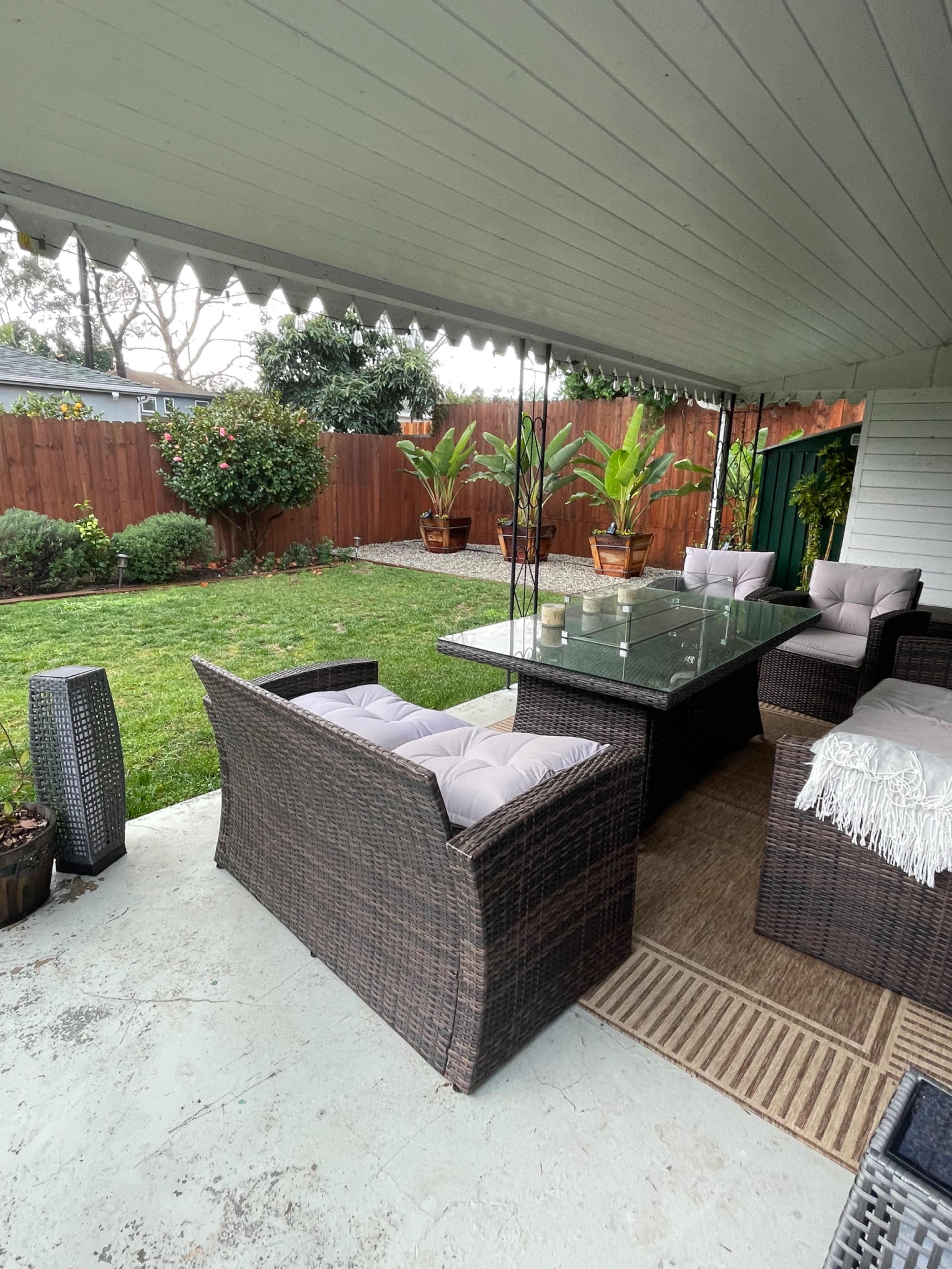 A covered patio features wicker furniture, a glass table, and planters with plants, overlooking a grassy backyard.