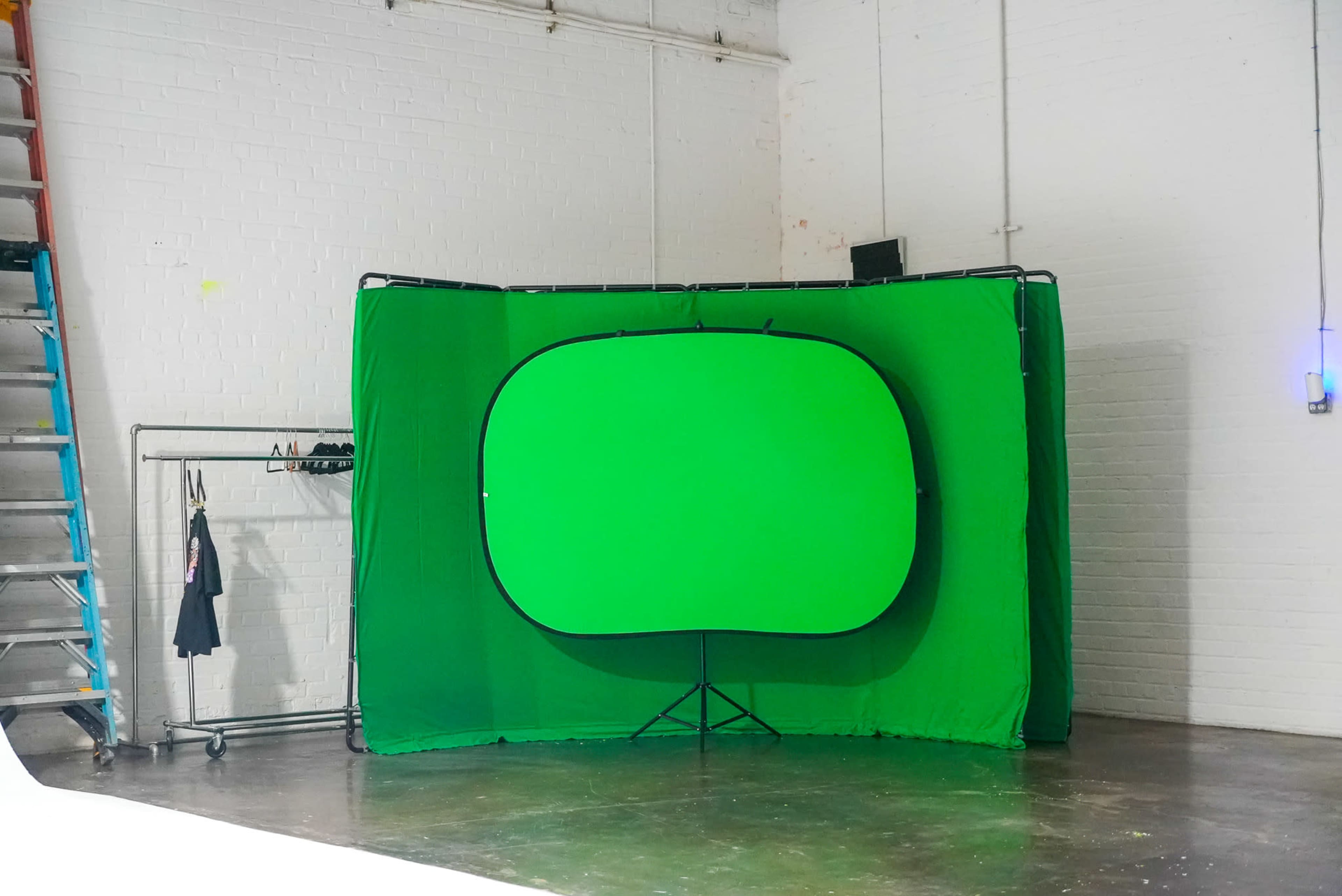 The image shows a green screen setup in a photography or video studio, with a folding frame and a ladder against a white wall.