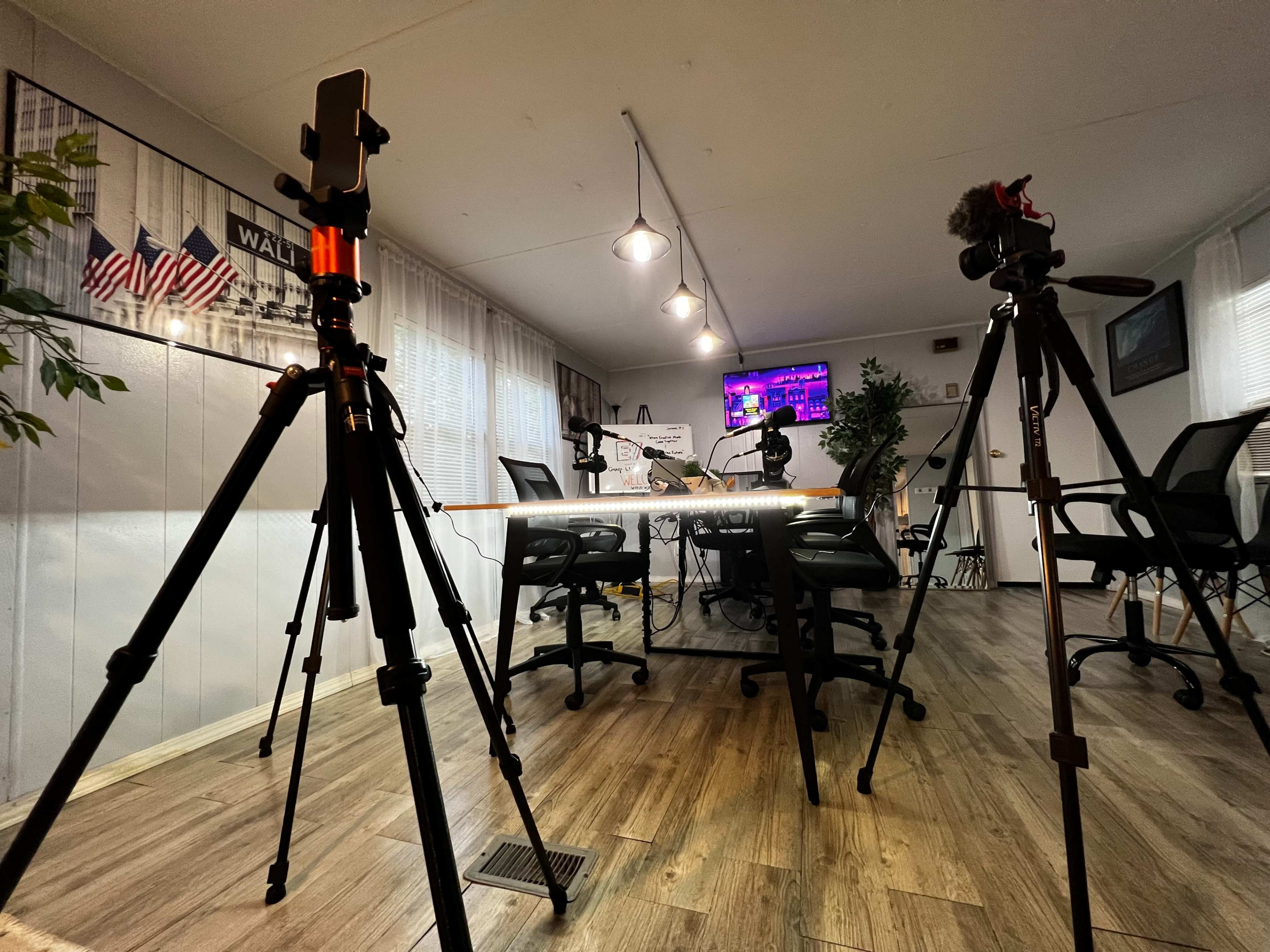 A brightly lit podcast recording studio equipped with tripods, microphones, and a large television screen displaying colorful graphics.