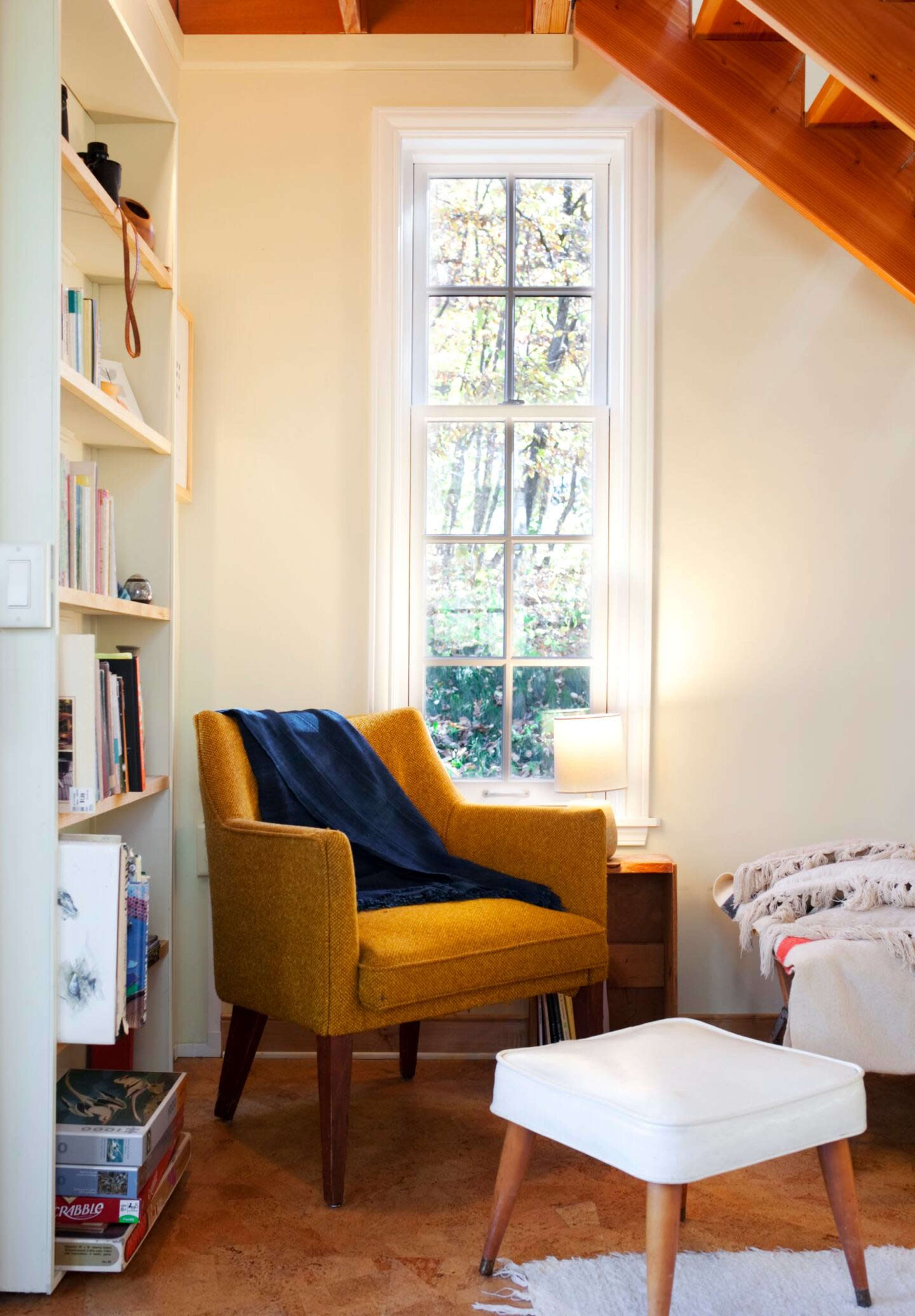 A cozy reading nook features a yellow armchair next to a window, with a small table lamp and a white ottoman.