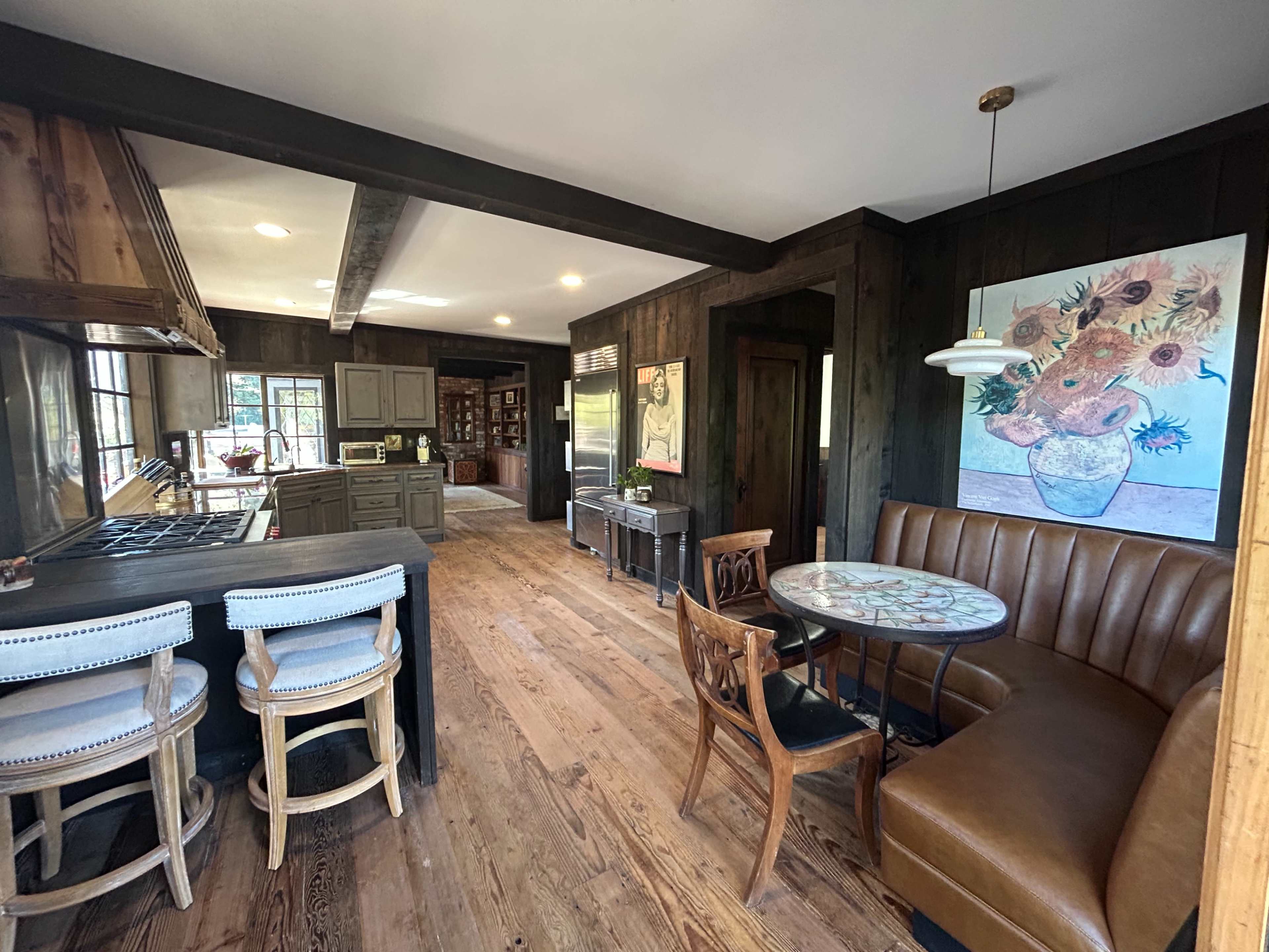 The image shows a rustic kitchen and dining area featuring a round table, a padded booth, and a bar with stools, all set against wooden walls and floors.