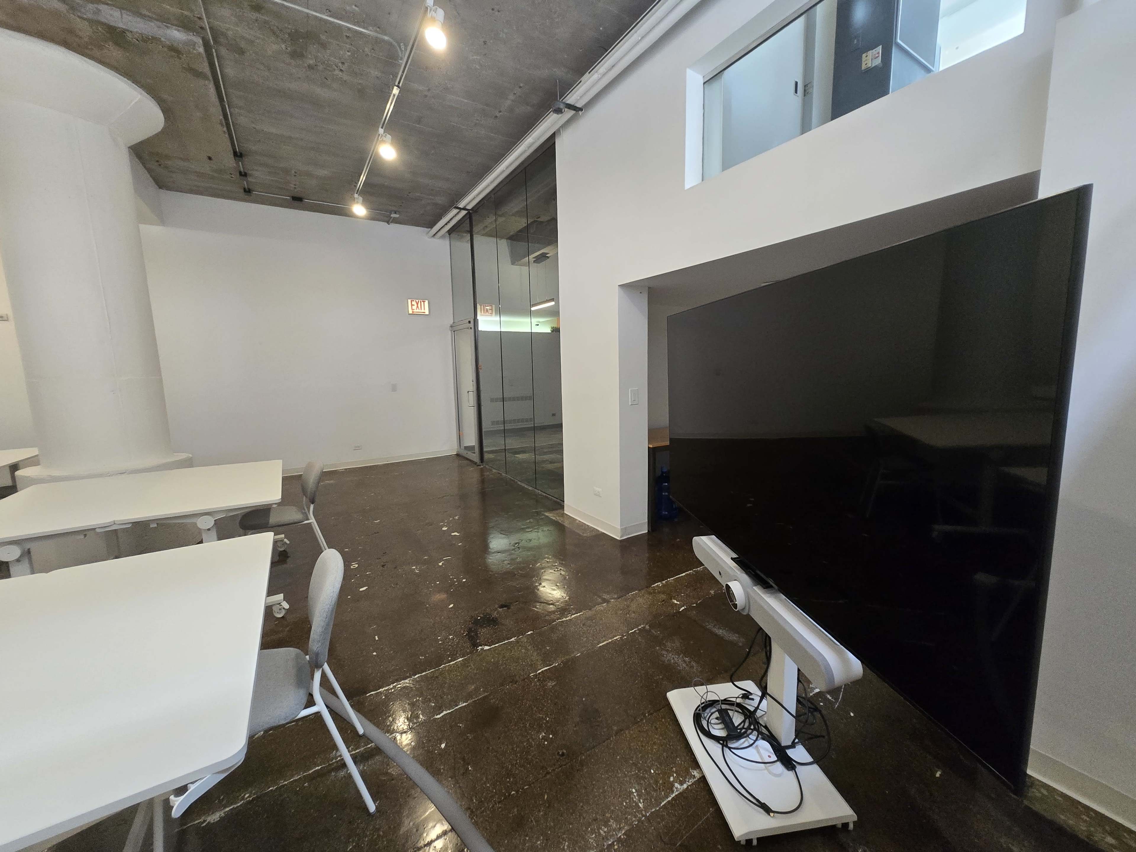 The image shows a modern office space featuring white walls, polished dark flooring, and a large screen on a stand, with tables and chairs arranged in the room.