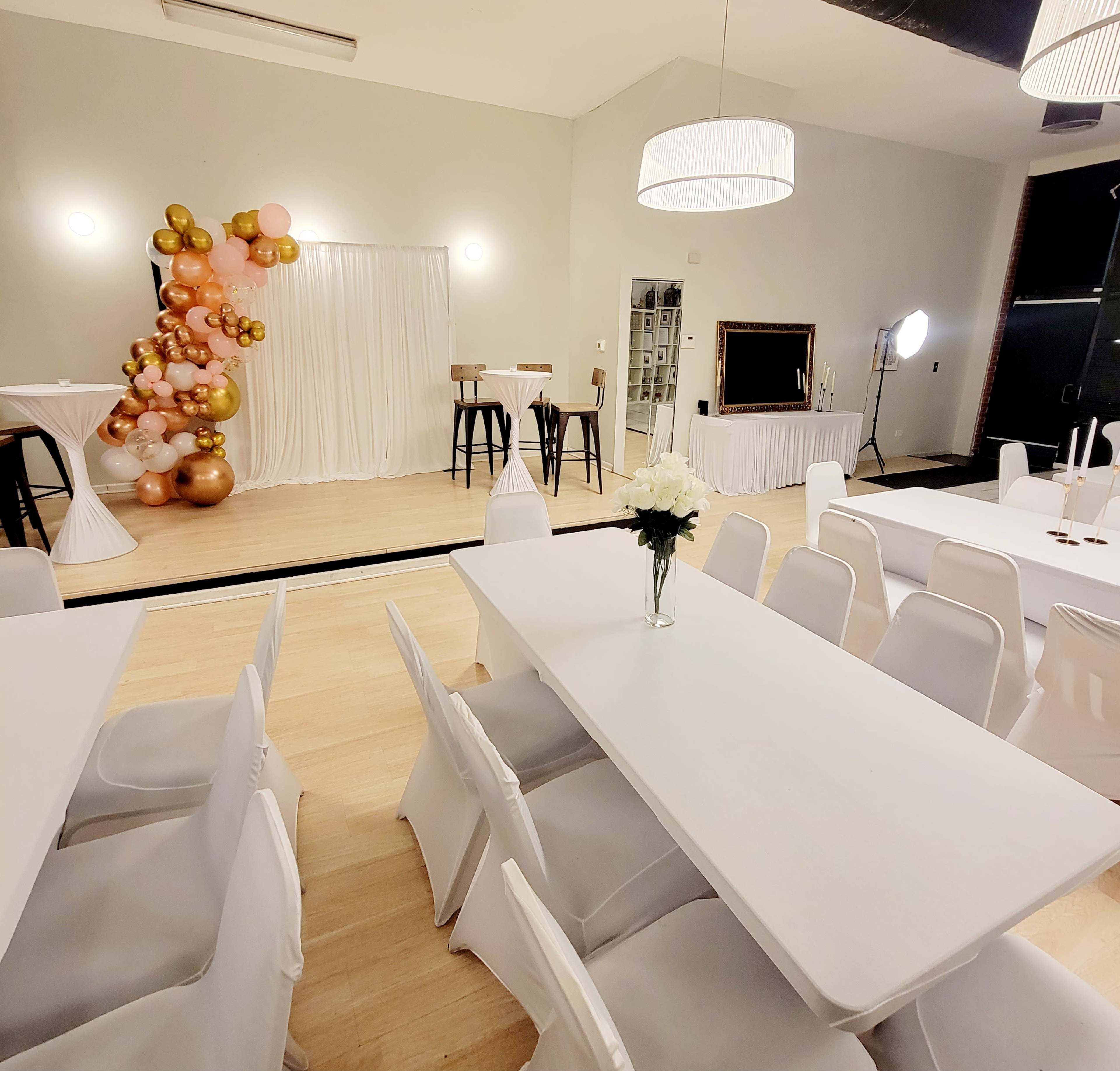 The image shows an elegantly set event space with white tables and chairs, a decorative backdrop of balloons, and a small stage with bar stools.
