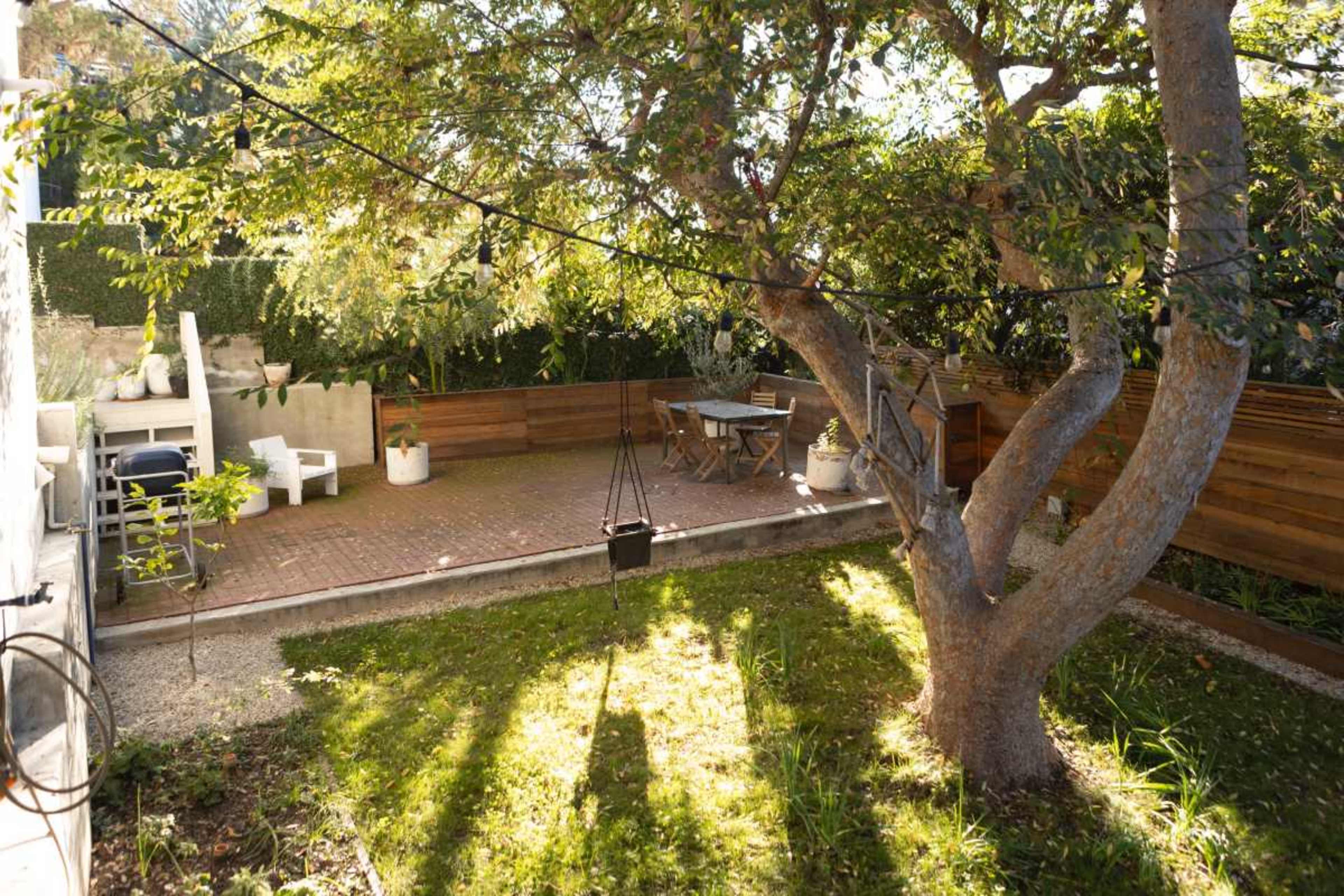The image shows a garden area with a large tree, a patio paved with bricks, and various outdoor furniture including a table and chairs.