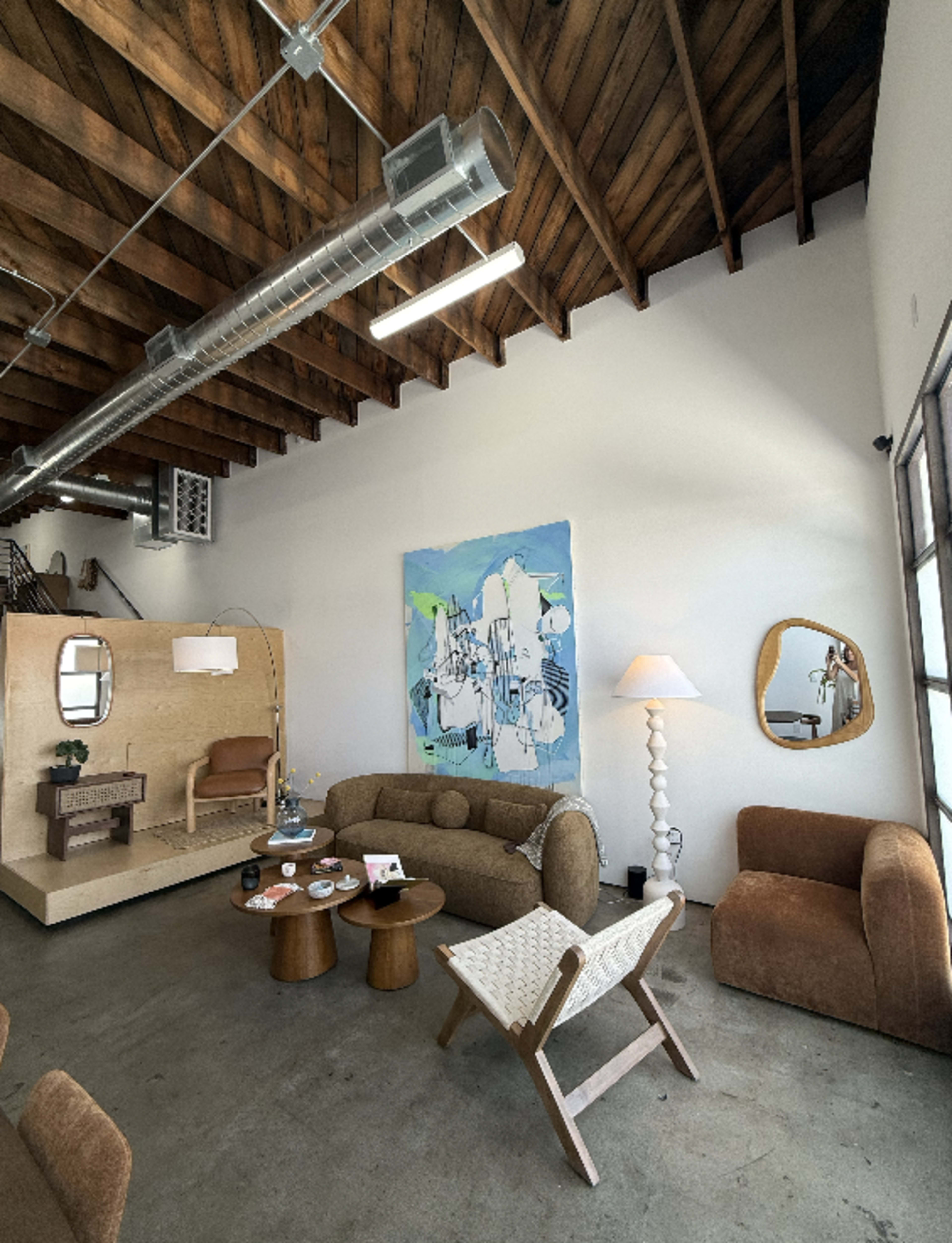 The image shows a modern interior living space featuring a curved brown sofa, a light wooden coffee table, an armchair, and a large abstract painting on the wall.