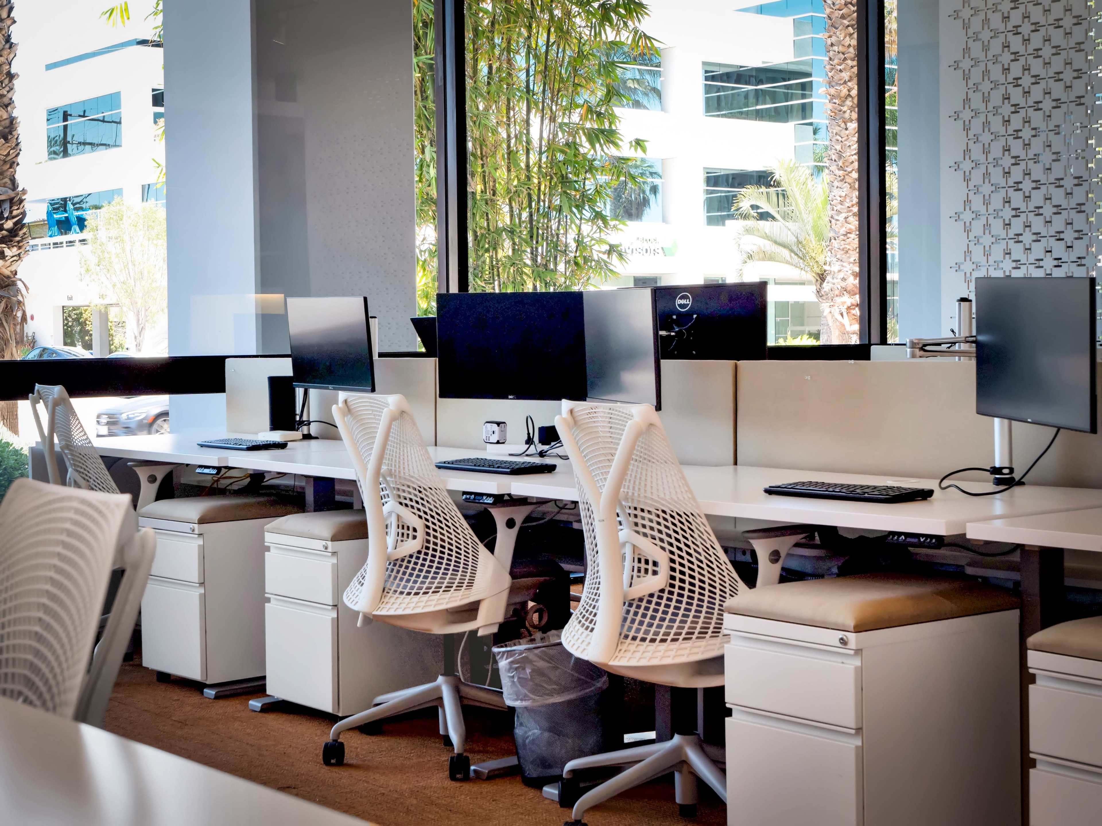 A modern office space with several white ergonomic chairs and computer desks near large windows.