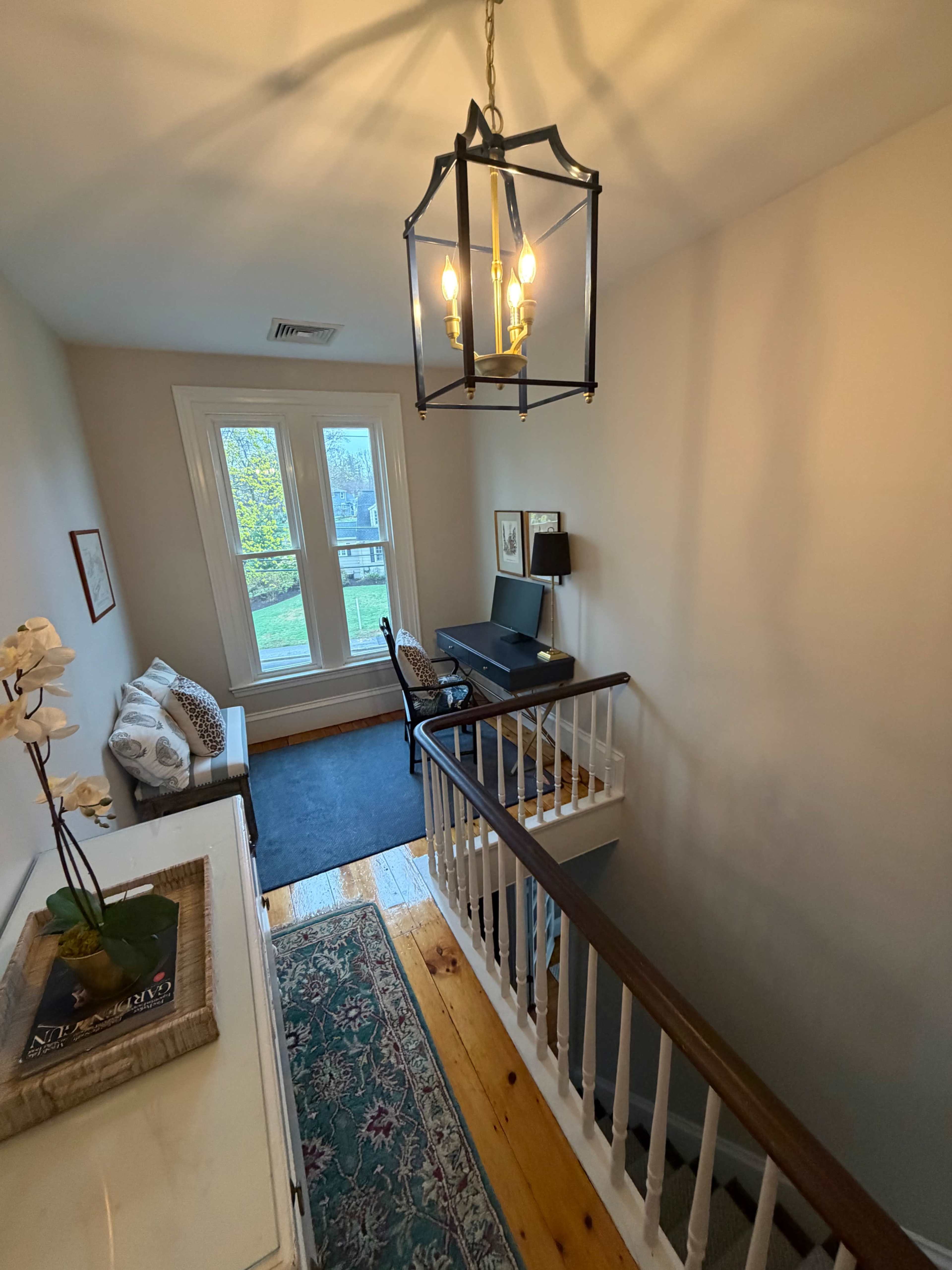 The image shows a well-lit staircase leading to a small landing area with a desk and chair, a sofa, and a window.