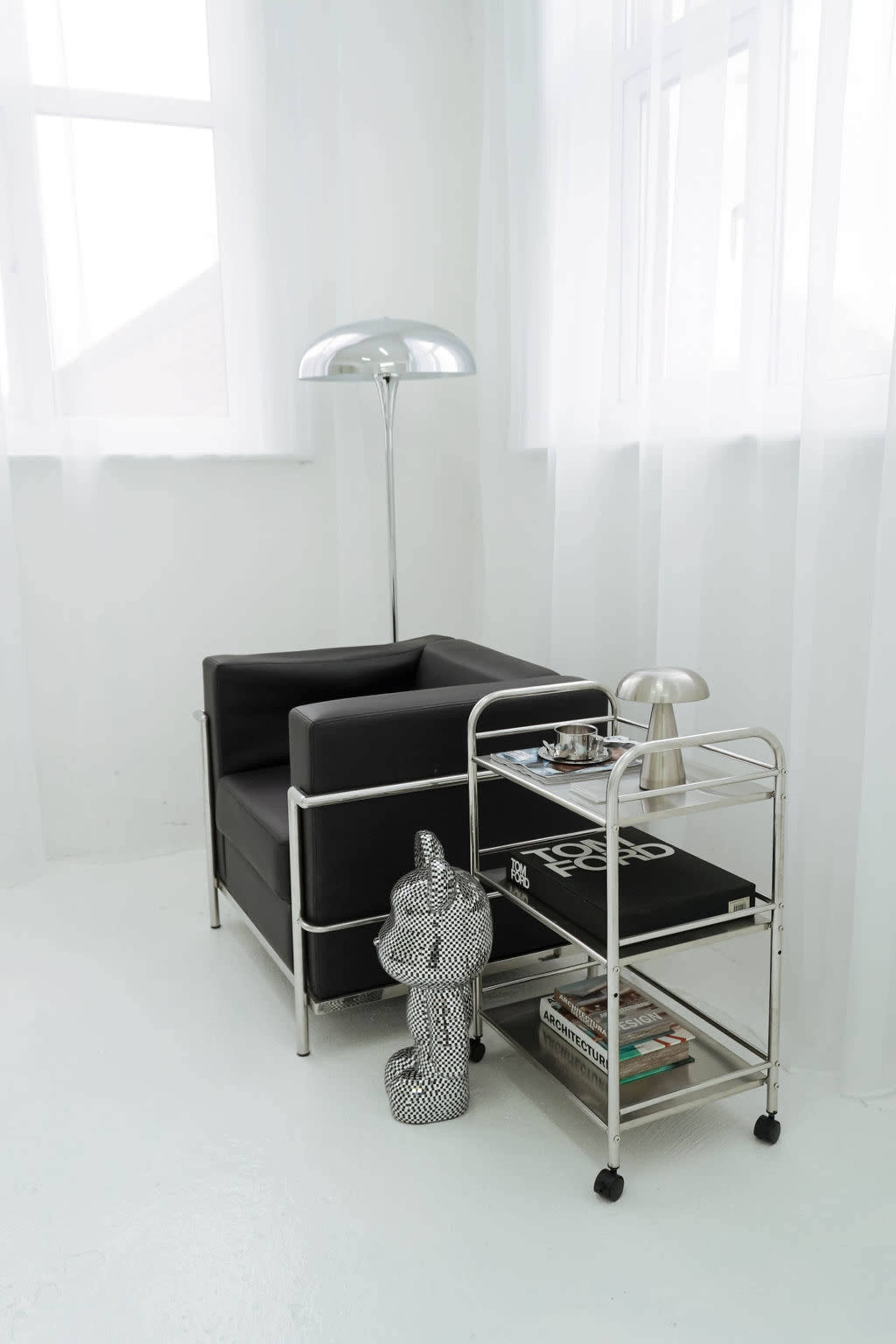 The image shows a minimalist living space featuring a black armchair, a metallic floor lamp, a silver lamp on a rolling cart, and a decorative statue.