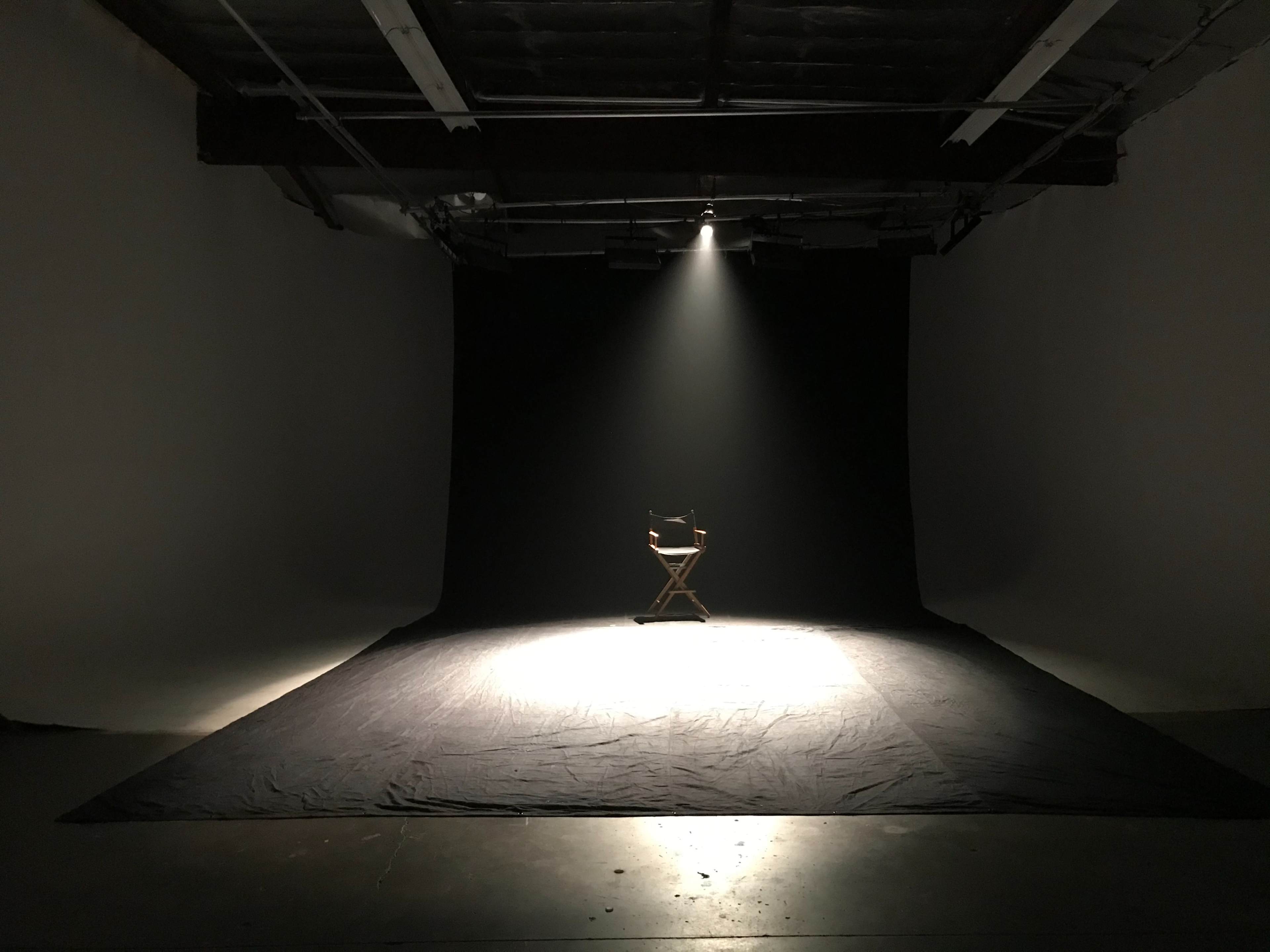 A solitary director's chair sits in a spotlight on a darkened stage surrounded by blank walls.