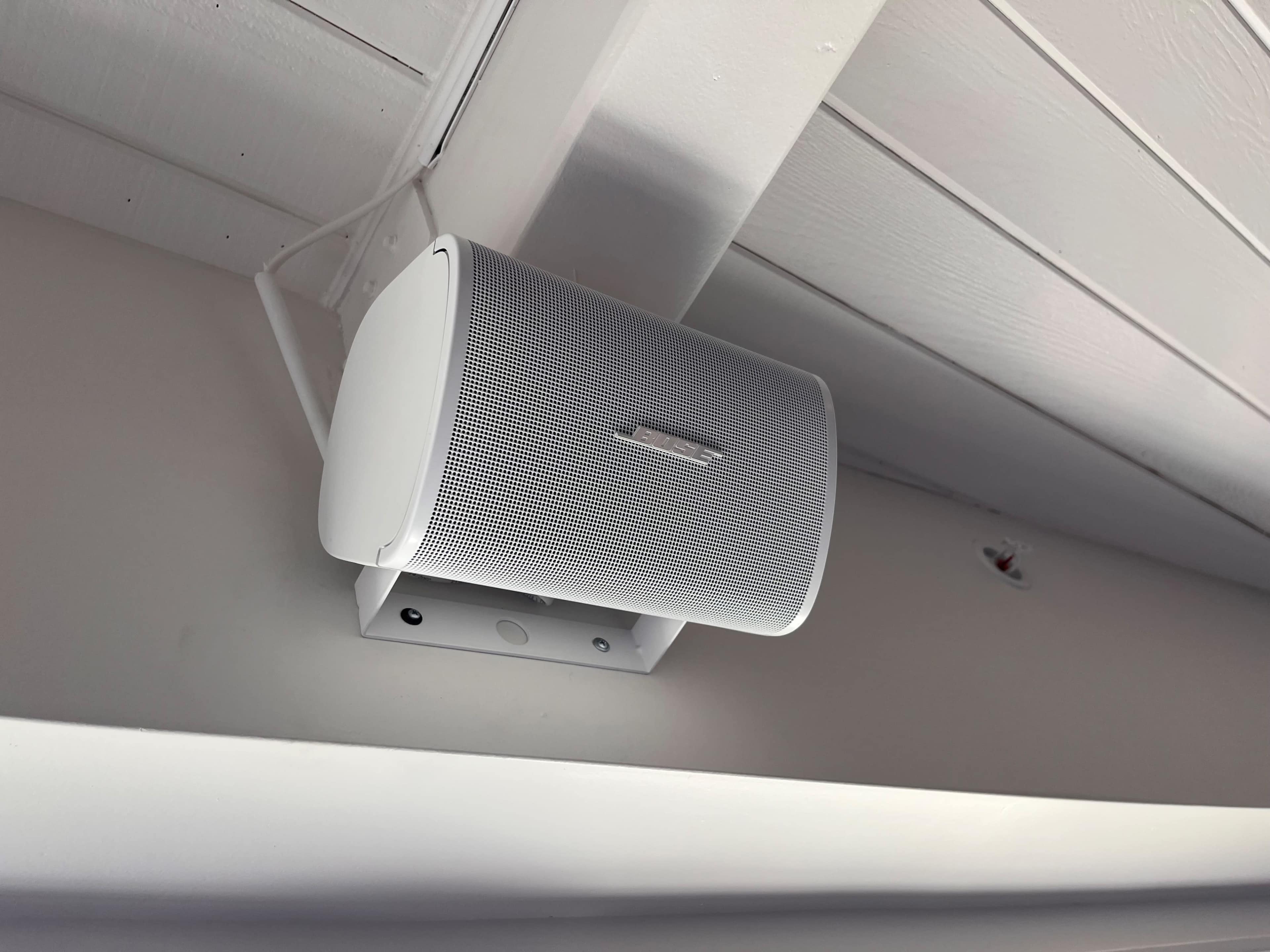 A white speaker is mounted on a wall beneath a wooden ceiling beam.