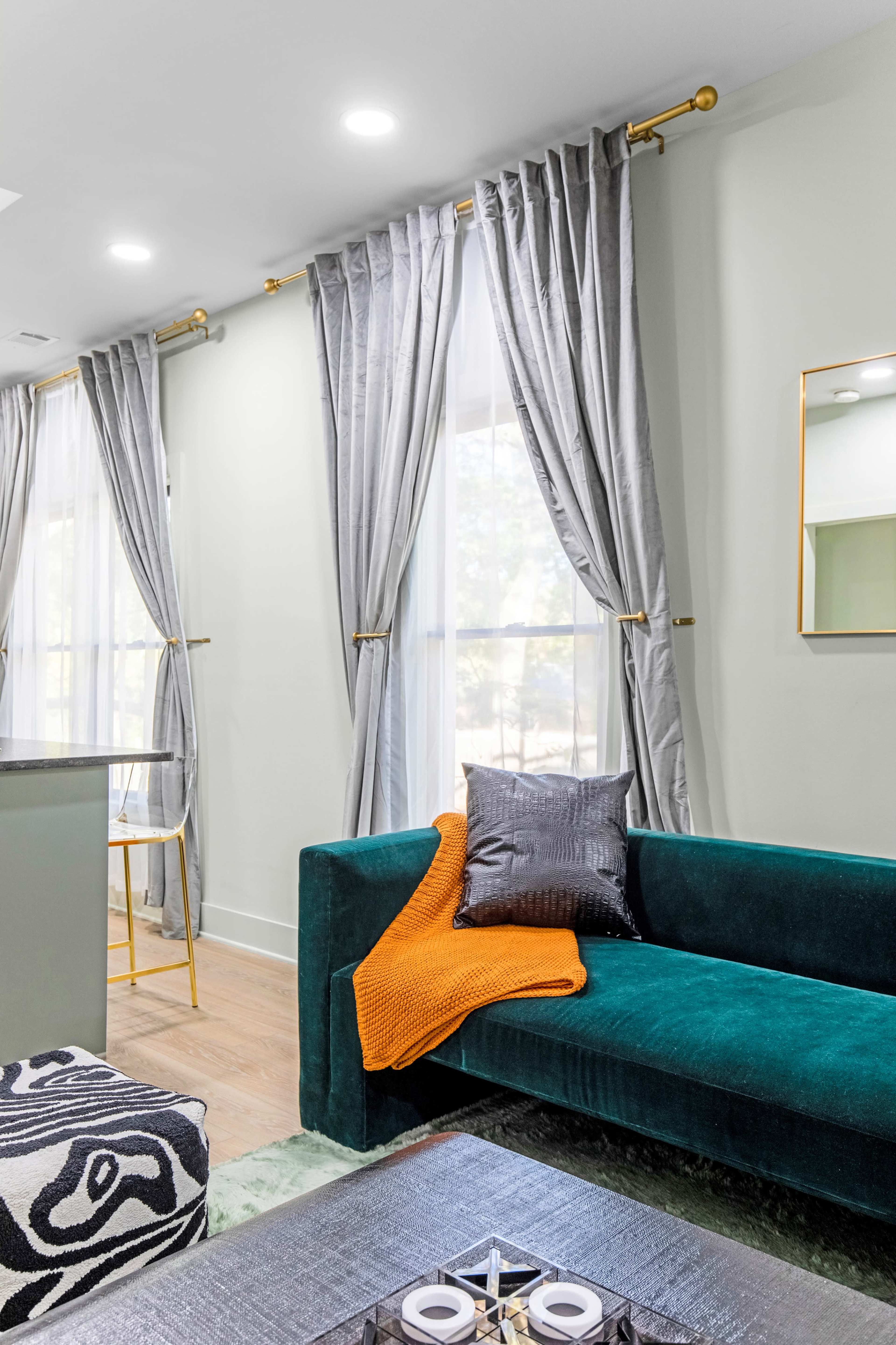 A modern living room features a teal sofa adorned with an orange throw and a gray pillow, alongside sheer gray curtains and a coffee table with decorative items.