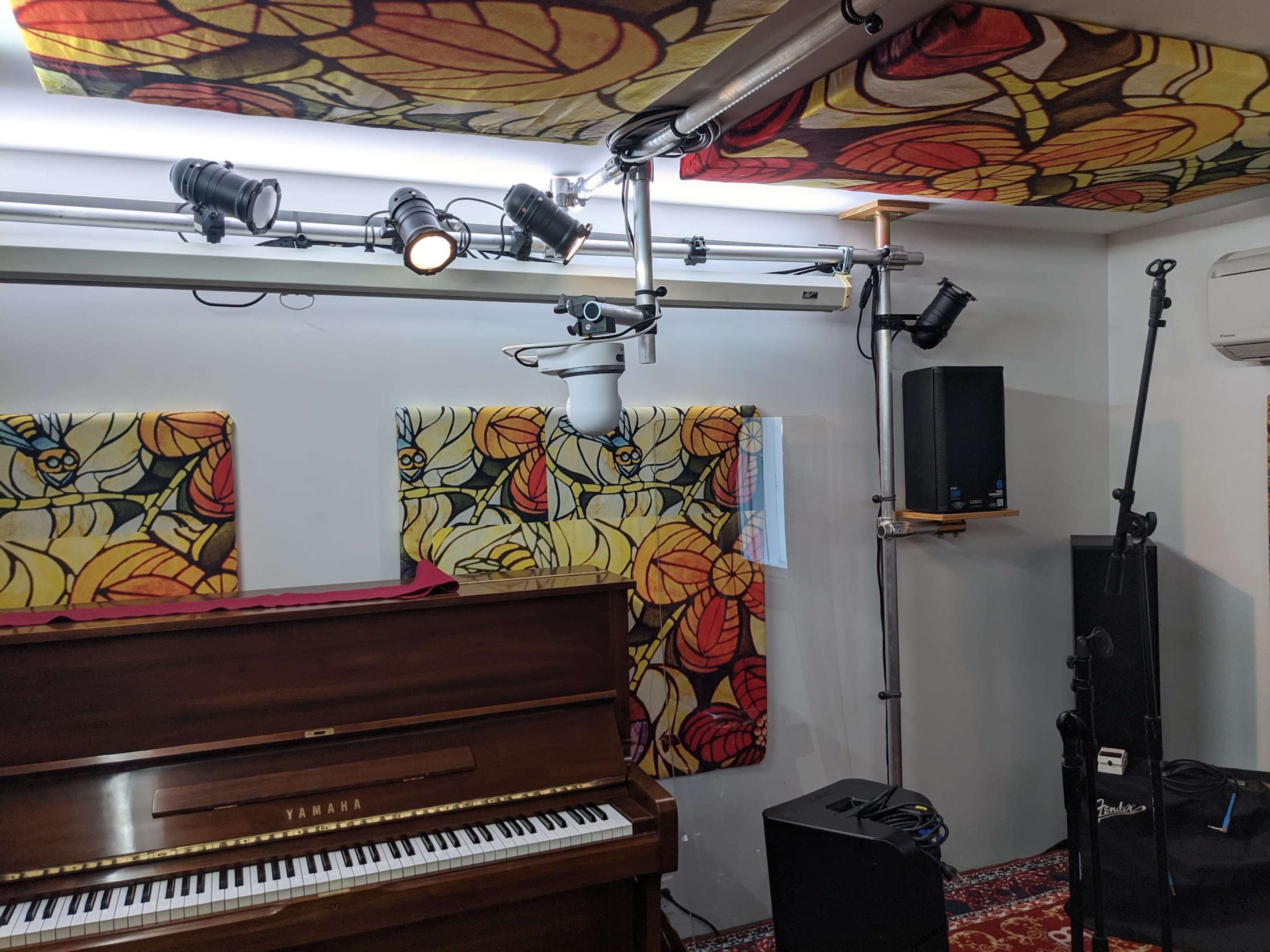 A music studio featuring a Yamaha piano, colorful floral-patterned acoustic panels, and various audio equipment including speakers and microphones.