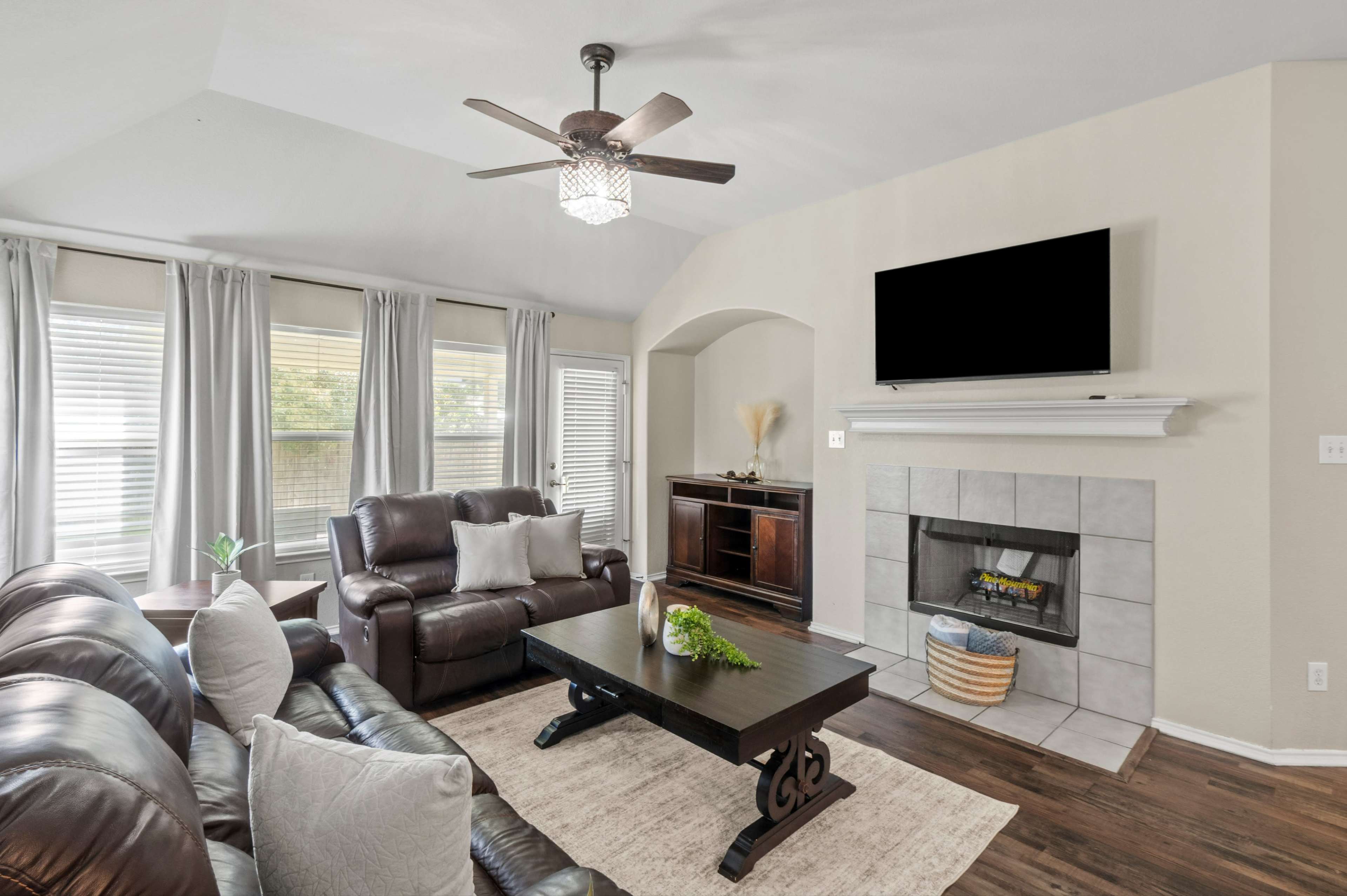 The living room features leather seating, a coffee table, a television mounted above a fireplace, and large windows with curtains.