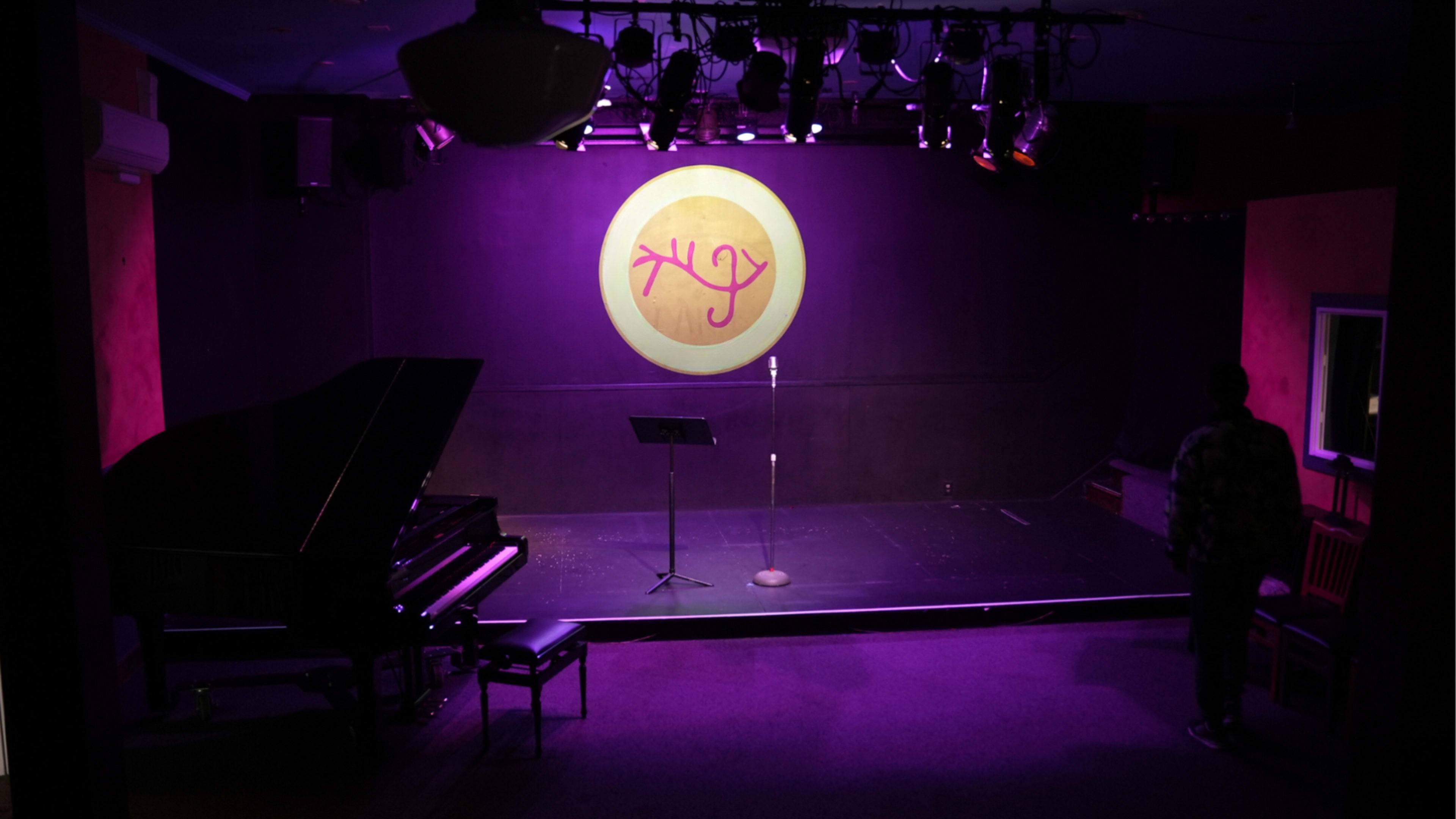 A dimly lit stage features a grand piano, a microphone stand, and a circular logo on the backdrop.