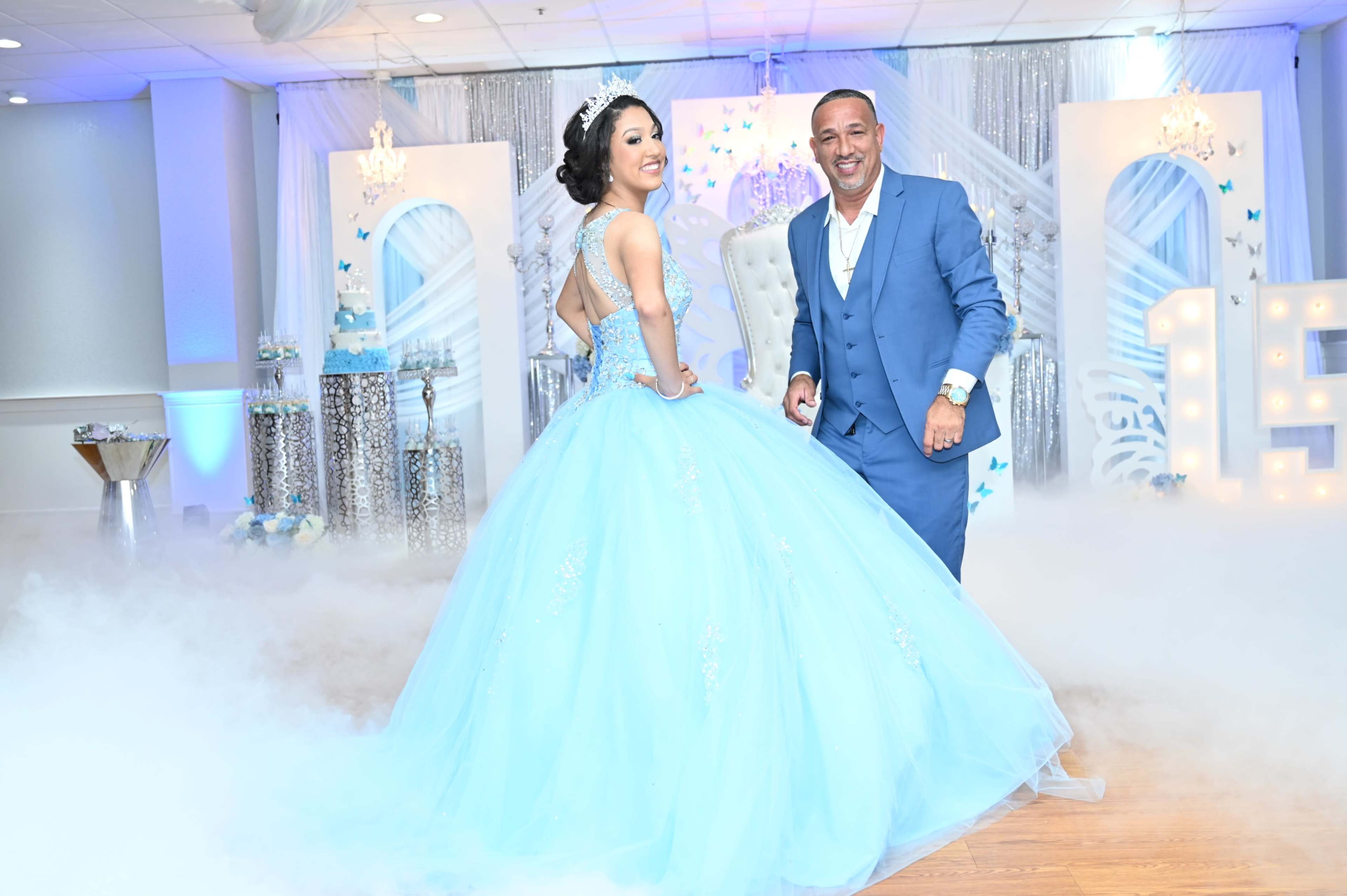 A young woman in a light blue ball gown poses with a man in a blue suit at a decorated event space with a misty floor.