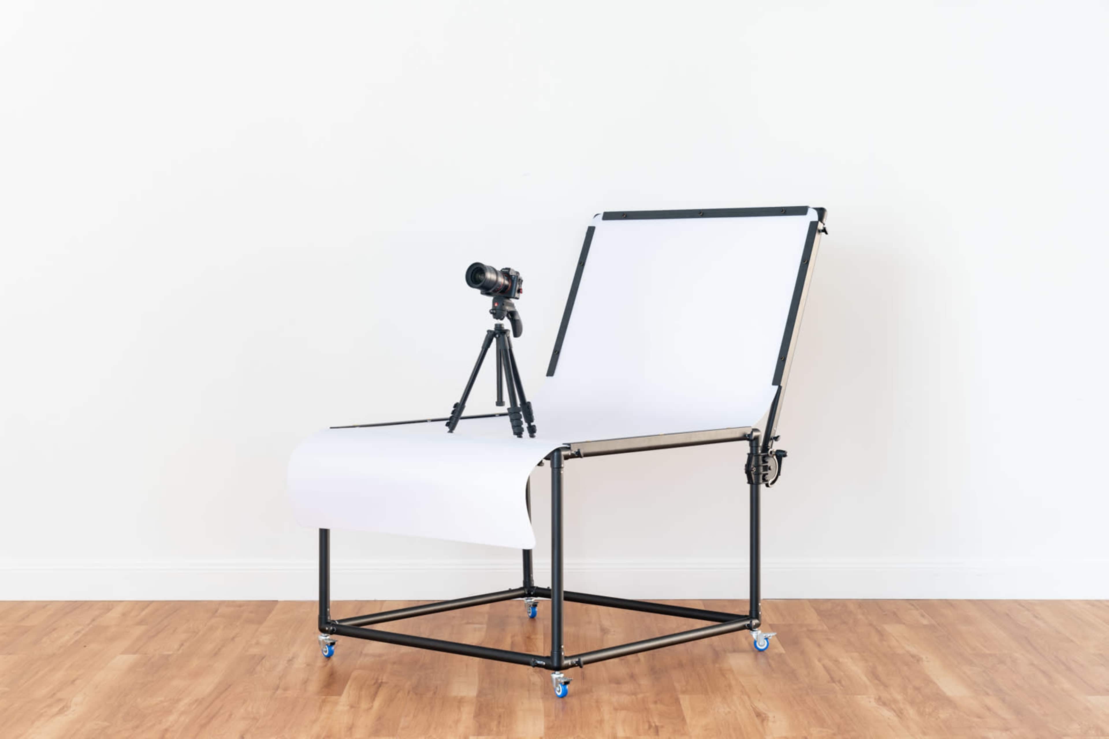 A photography setup featuring a camera on a tripod next to a large white backdrop on a mobile stand.