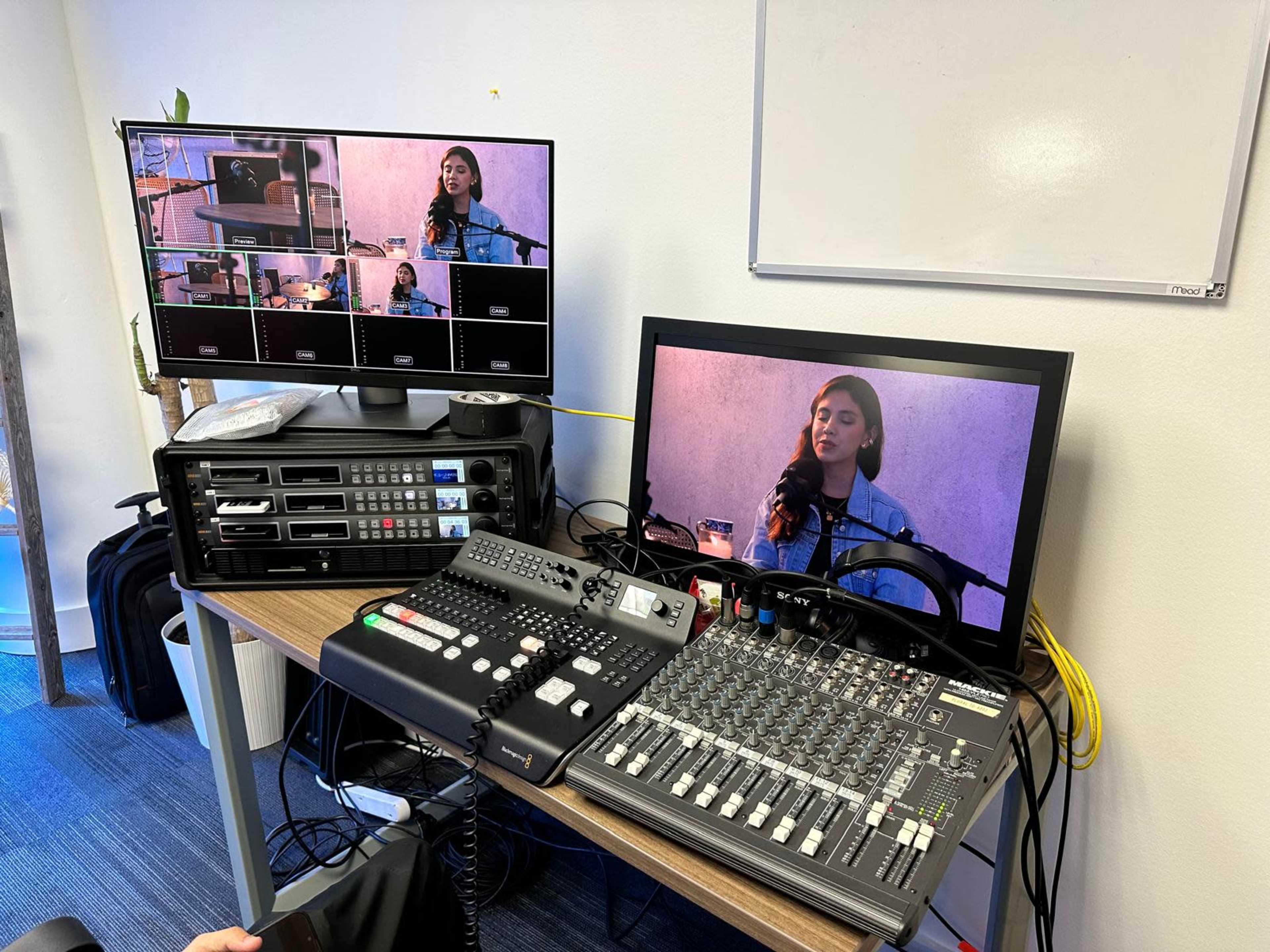 The image shows a video production setup with multiple monitors displaying a woman speaking into a microphone, along with a mixing console and various audio/video equipment on a table.