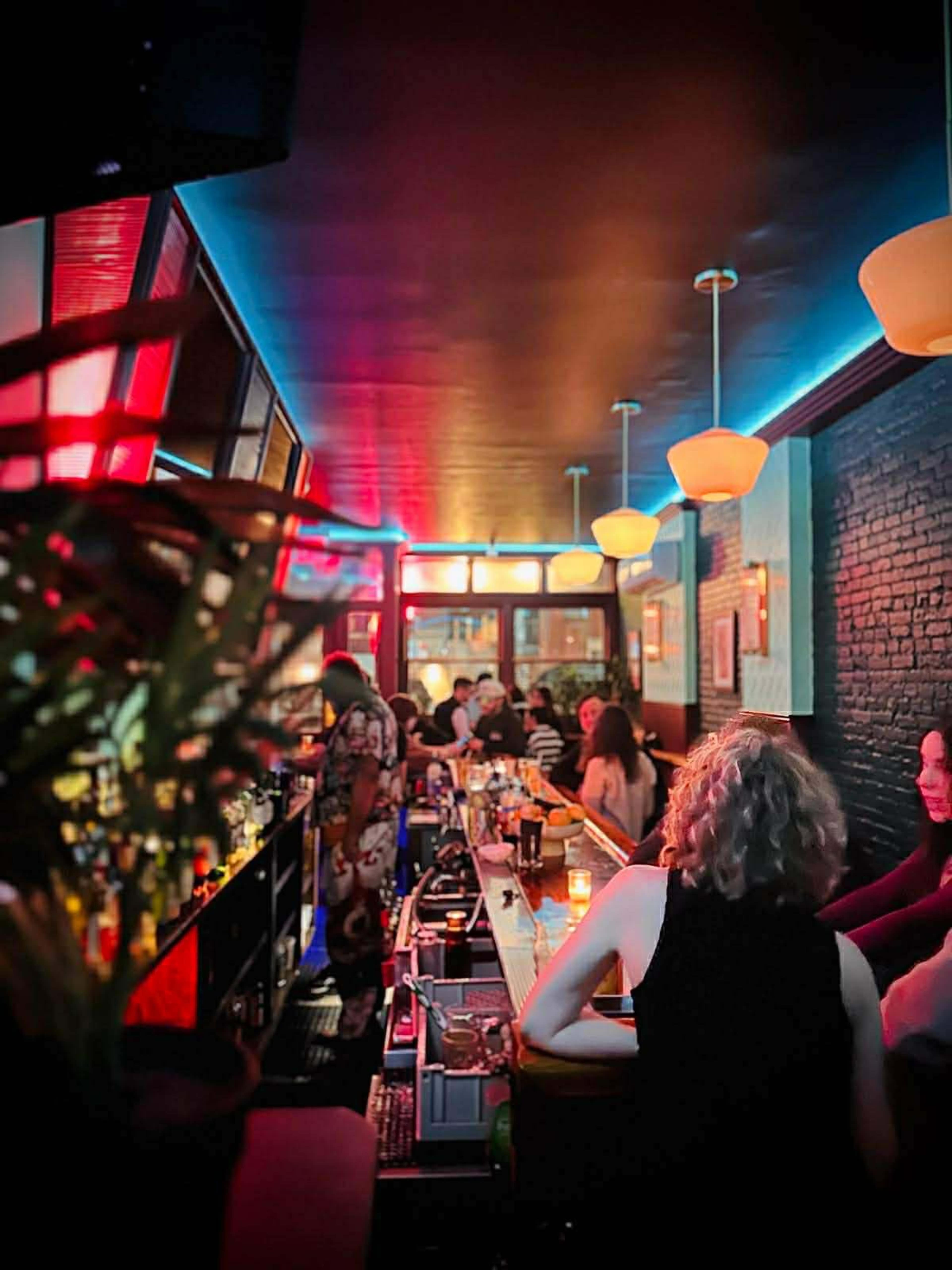 A narrow bar is filled with patrons, illuminated by colorful lights and lined with bottles along the back.