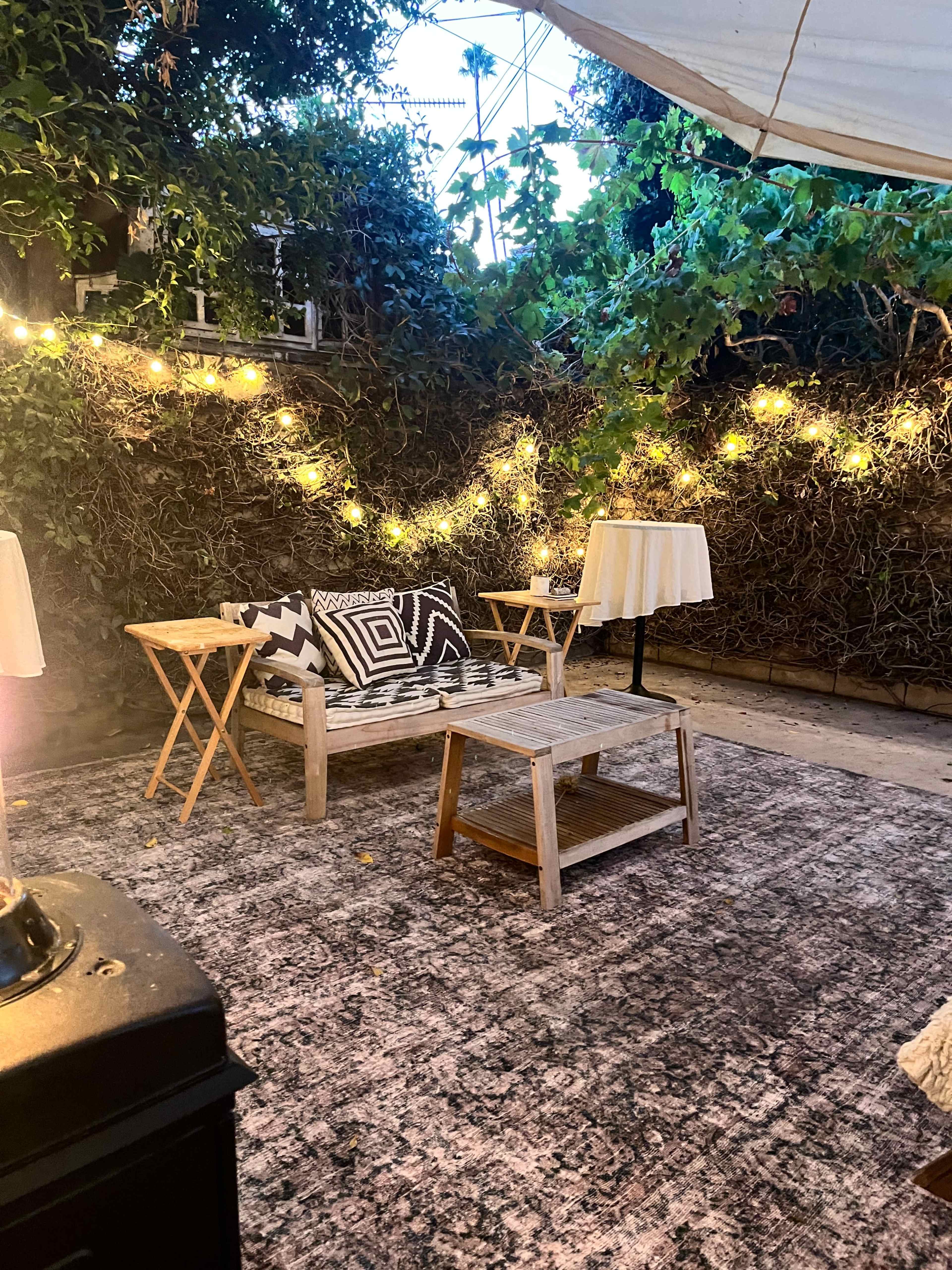 The image shows a cozy outdoor seating area with a wooden sofa and two folding tables, surrounded by lush green vines and strings of warm lights.
