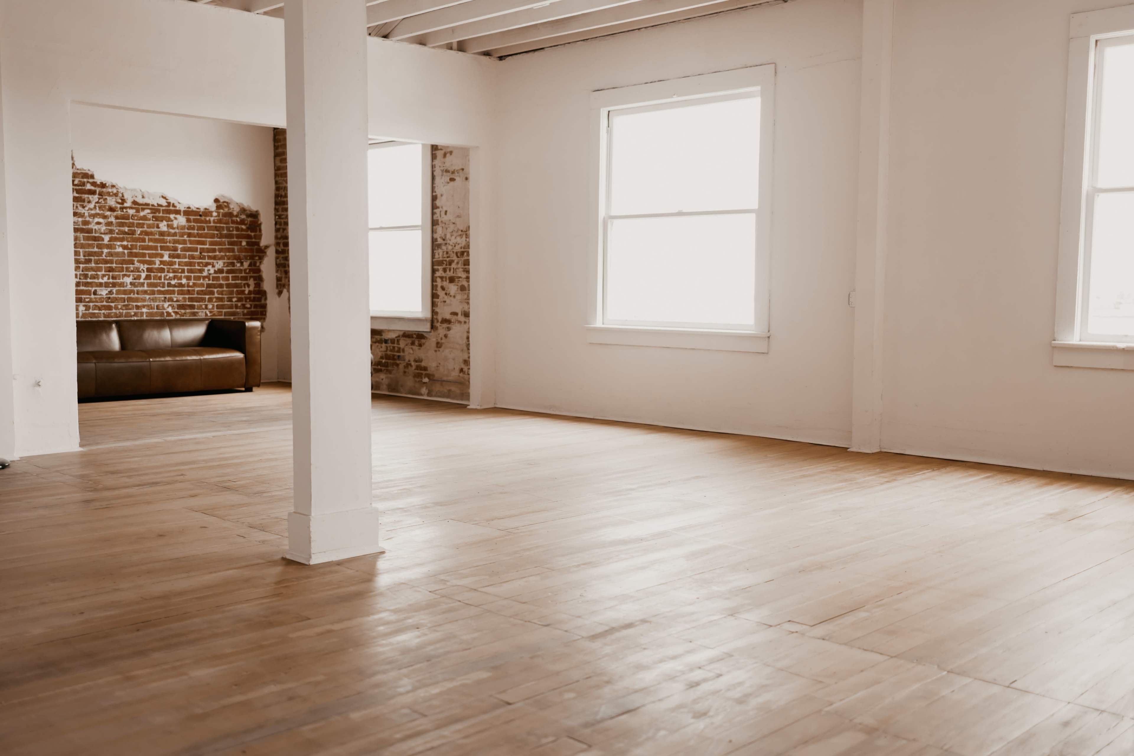 A spacious, empty room with wooden floors, bright windows, and exposed brick walls in one corner.