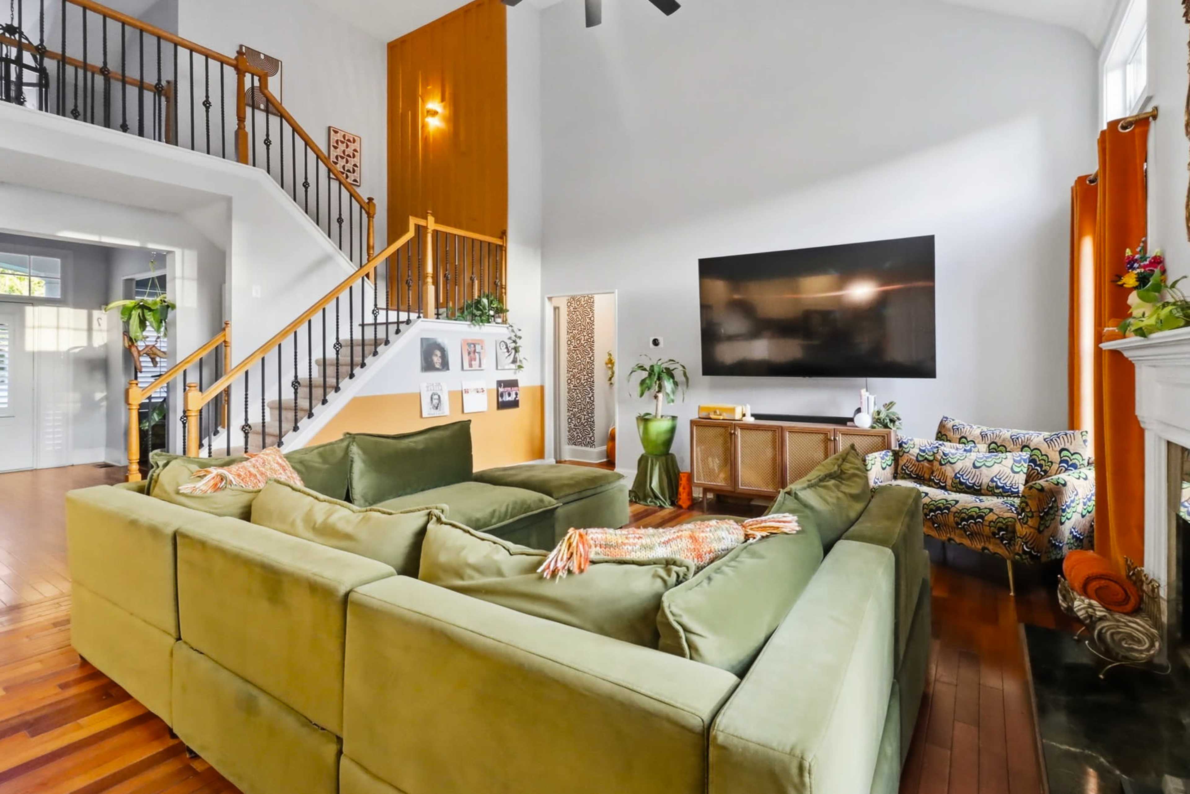 The image shows a spacious living room featuring a large green sectional sofa, a wooden staircase, and a wall-mounted television, with various decorative elements and furniture.