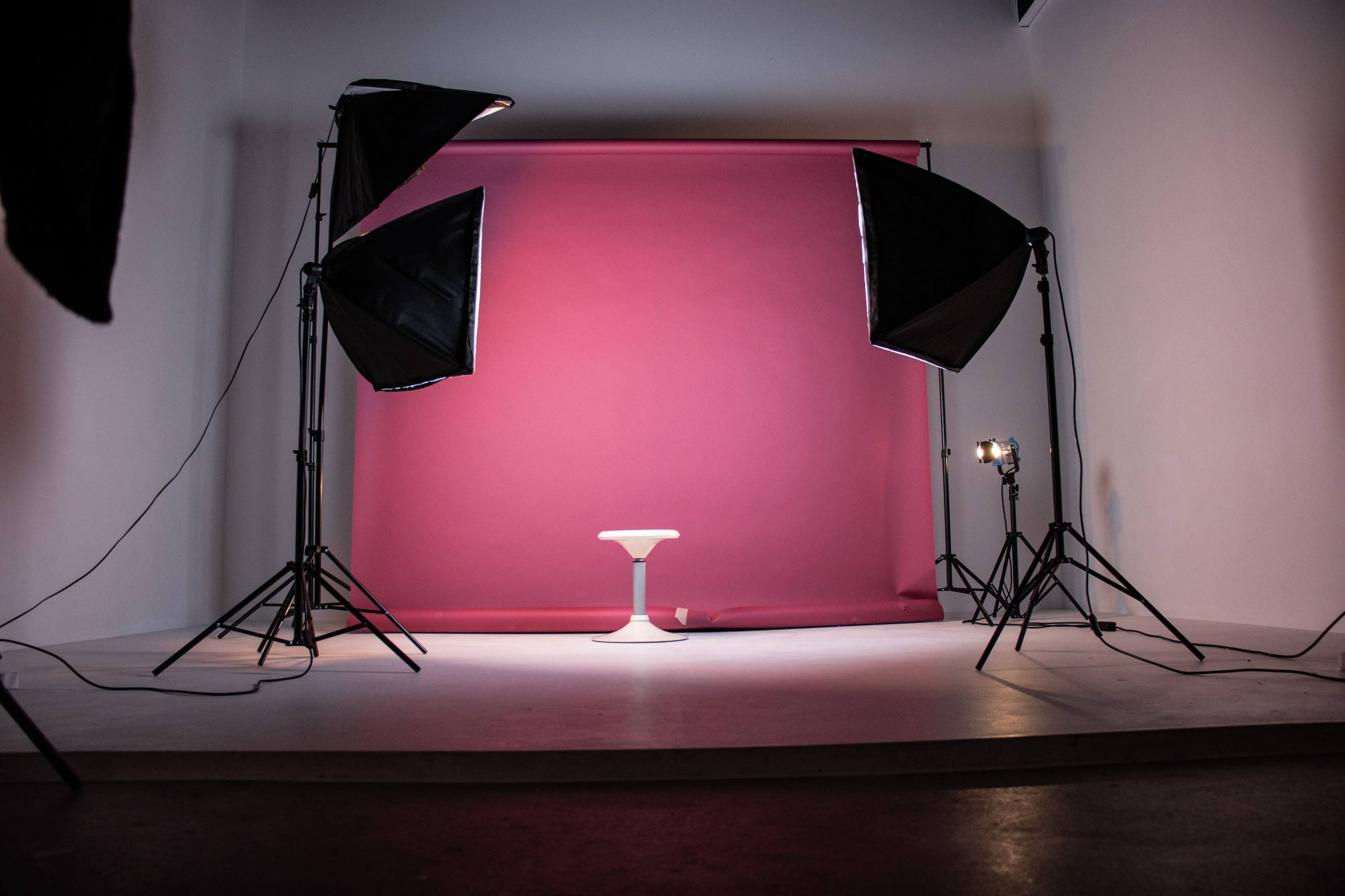 A photography studio setup features a pink backdrop with a white stool illuminated by softbox lights.