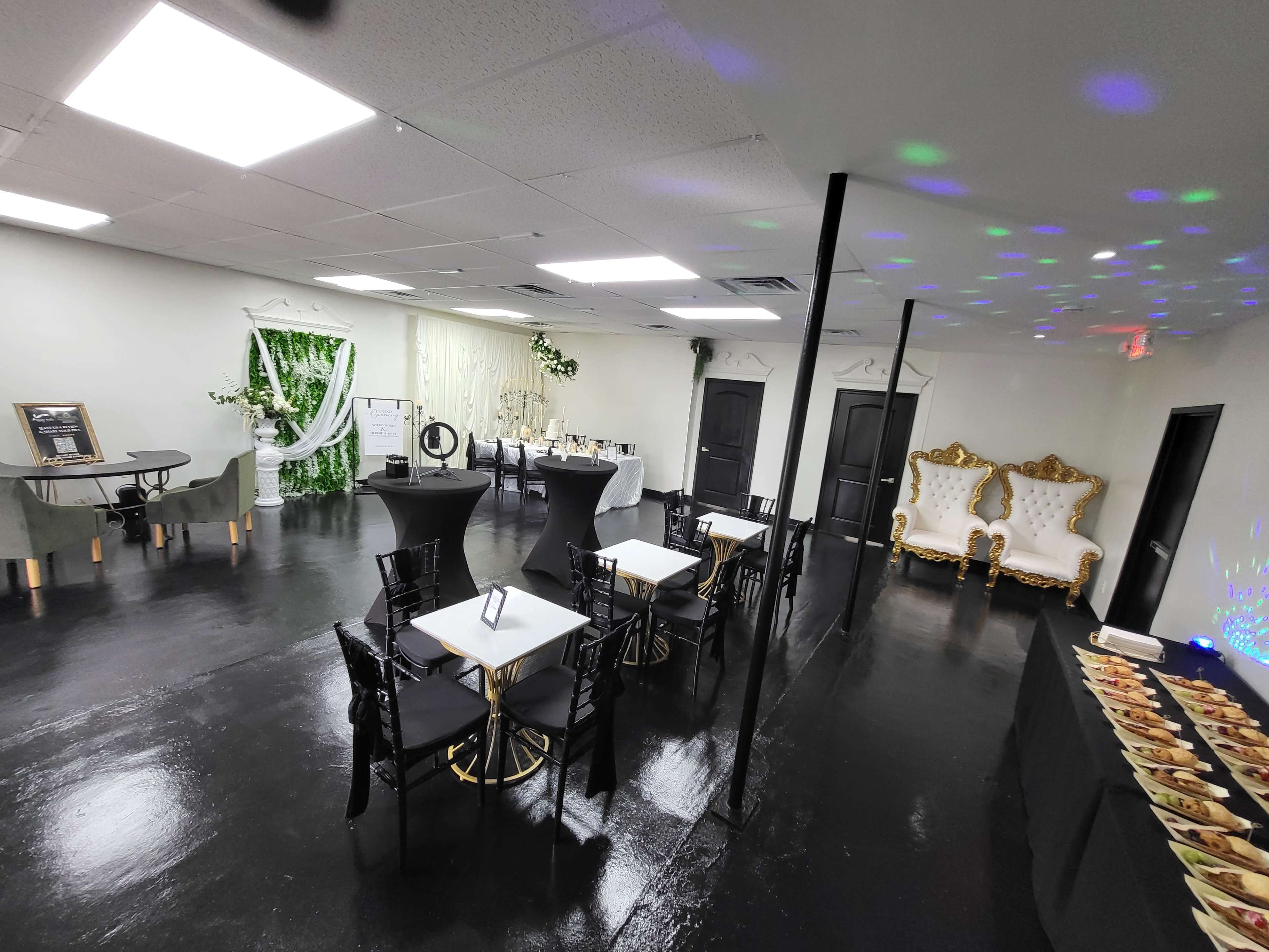 The image shows an elegantly arranged event space with tables, chairs, and decorative elements, featuring two ornate white chairs against a backdrop of greenery and ambient lighting.