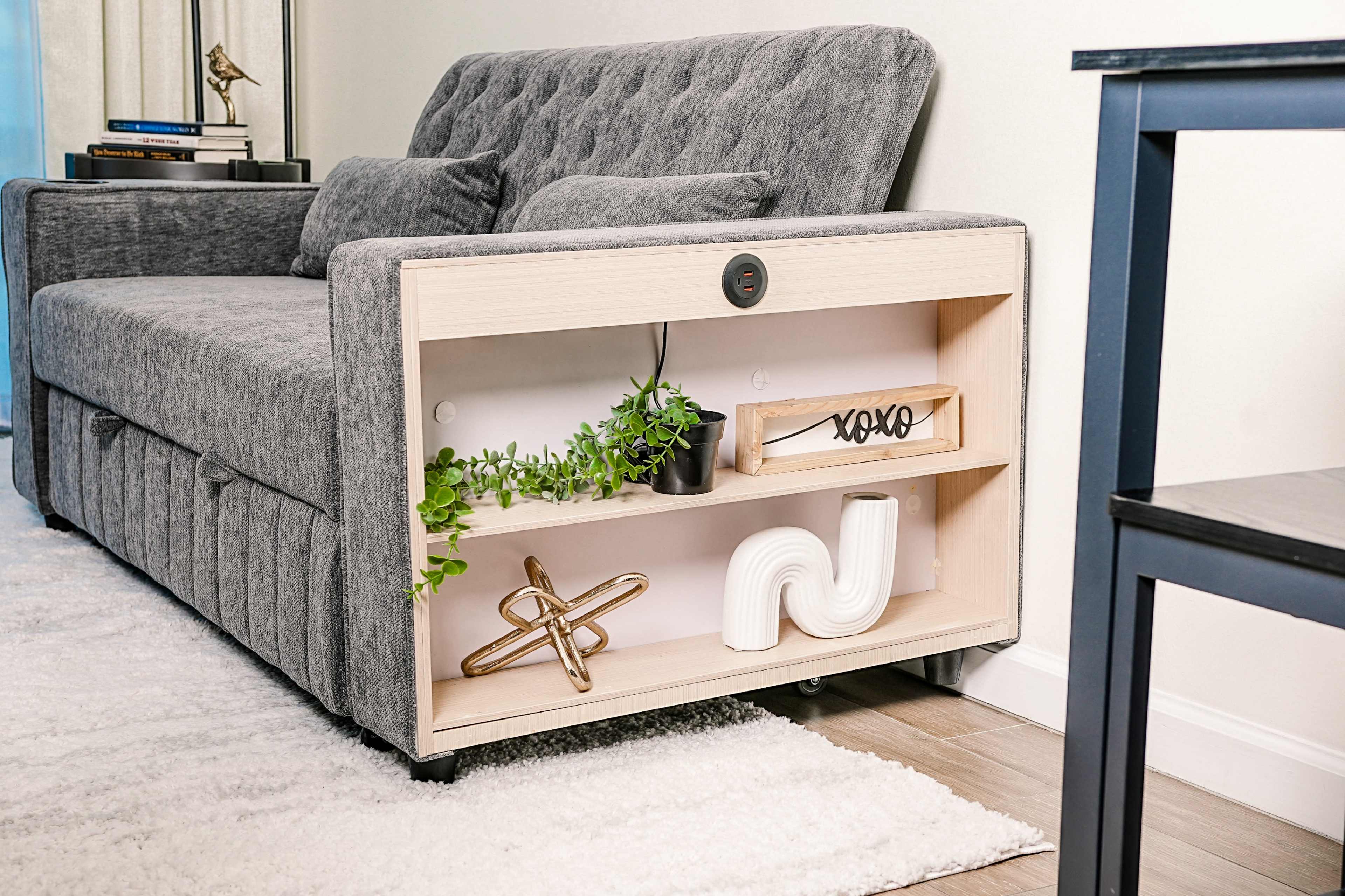 The image shows a modern gray sofa with a wooden shelf that holds decorative items and plants, positioned next to a small black side table on a light rug.