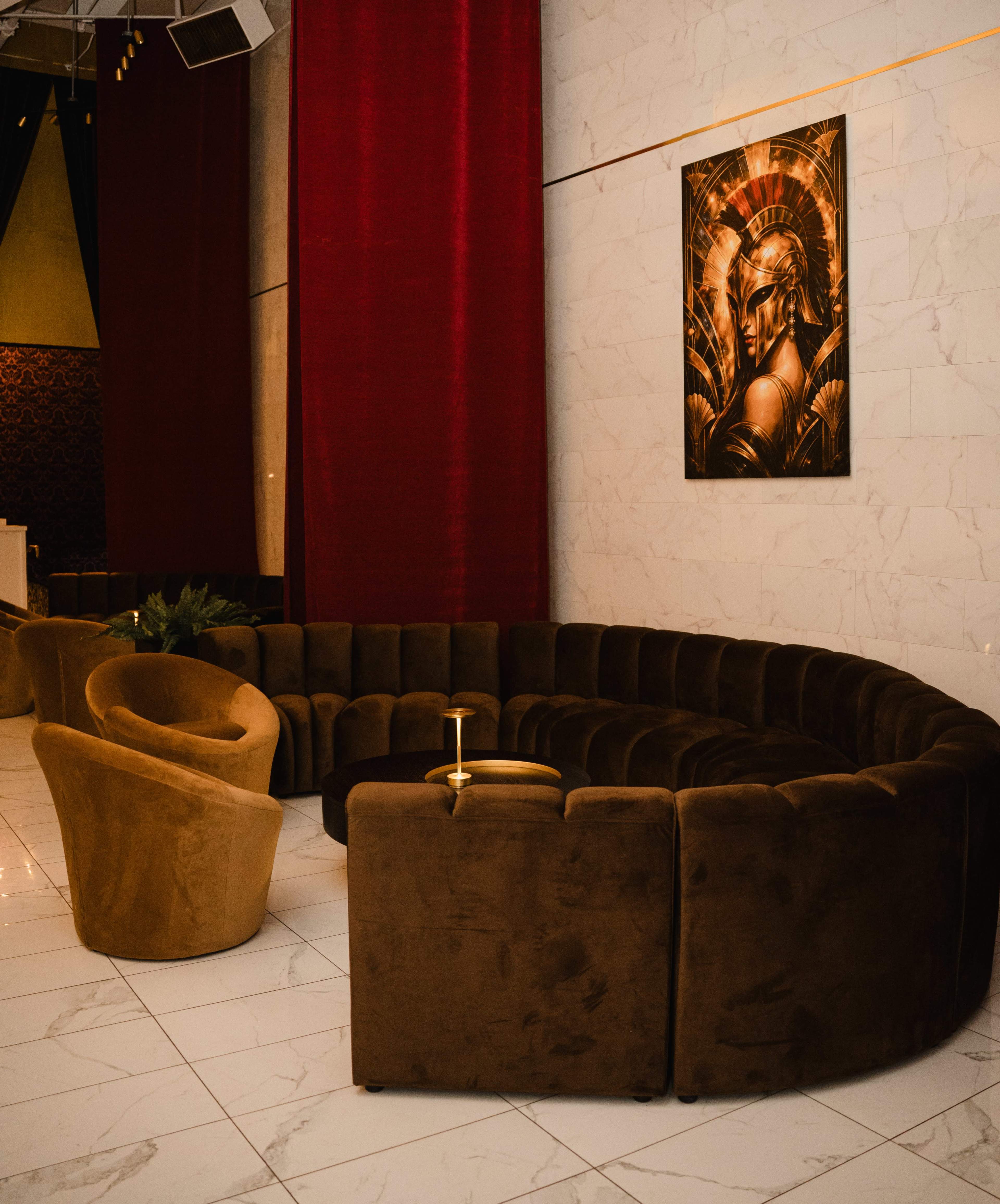 The image shows a circular brown couch set against a backdrop of red drapes and a marble wall, with a golden artwork depicting a figure on the wall above.