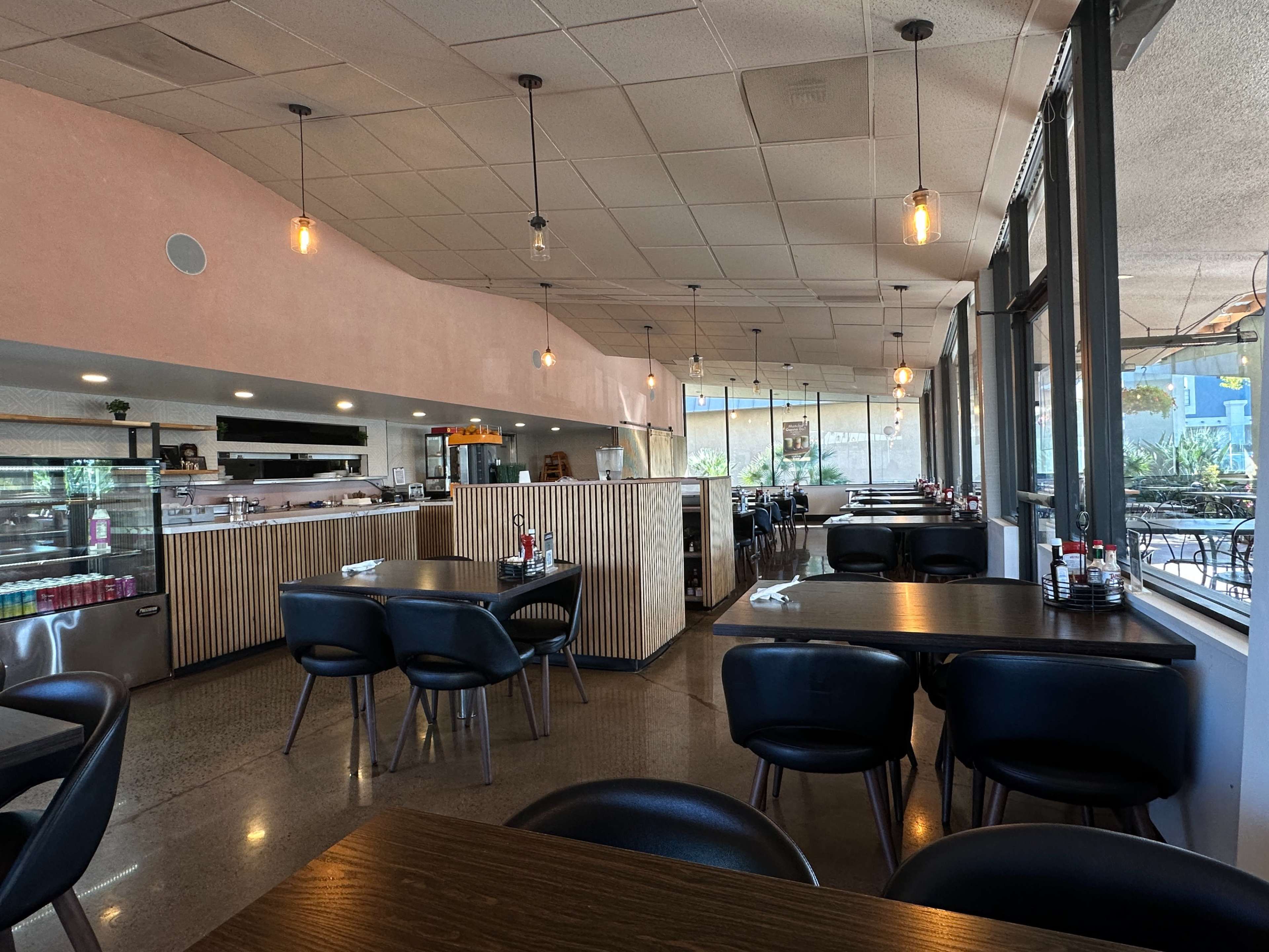 The image shows a modern restaurant interior with wooden tables, black chairs, and large windows allowing natural light to enter.