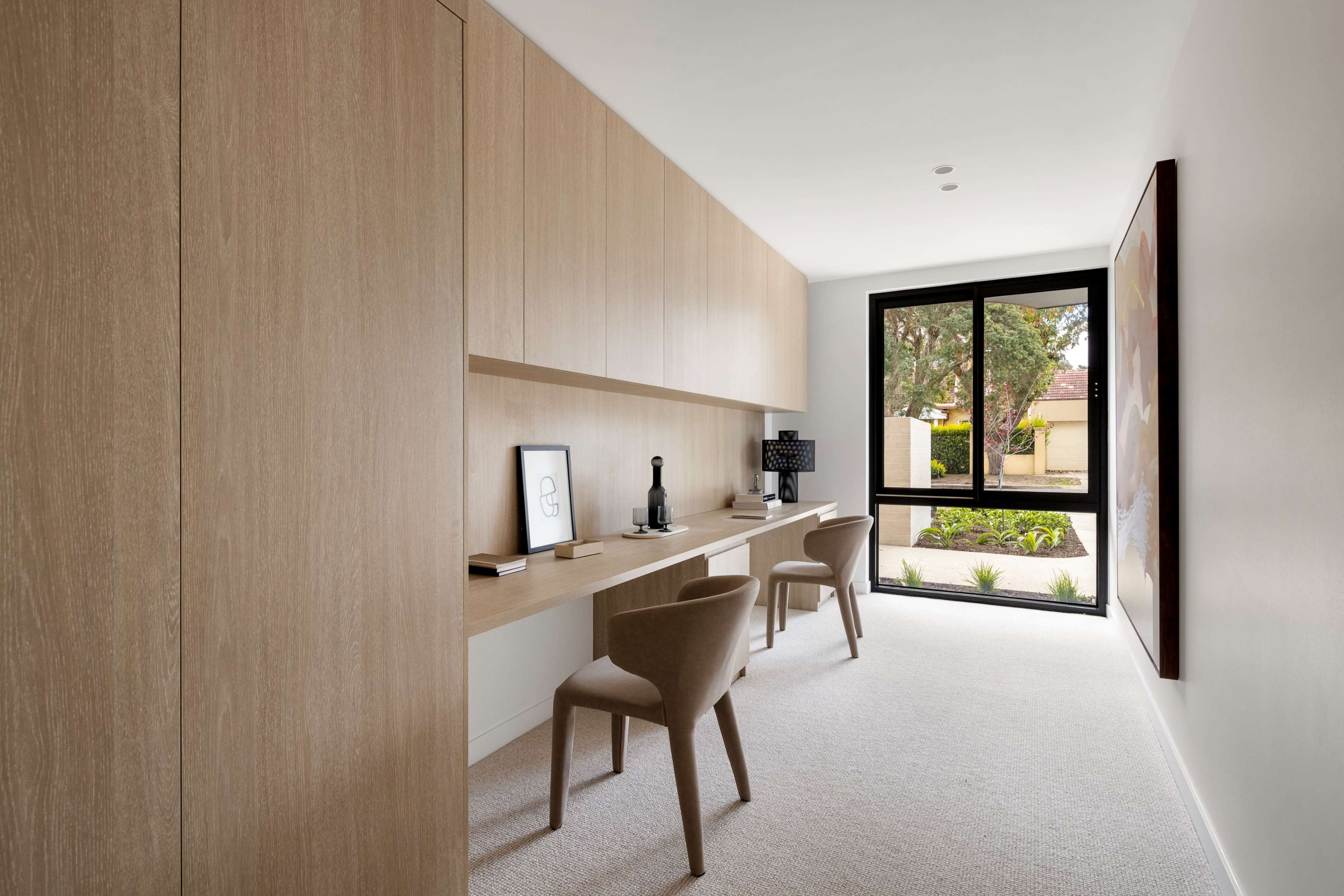 The image shows a modern home office with light wood cabinetry, a desk, two chairs, and large windows overlooking a garden.