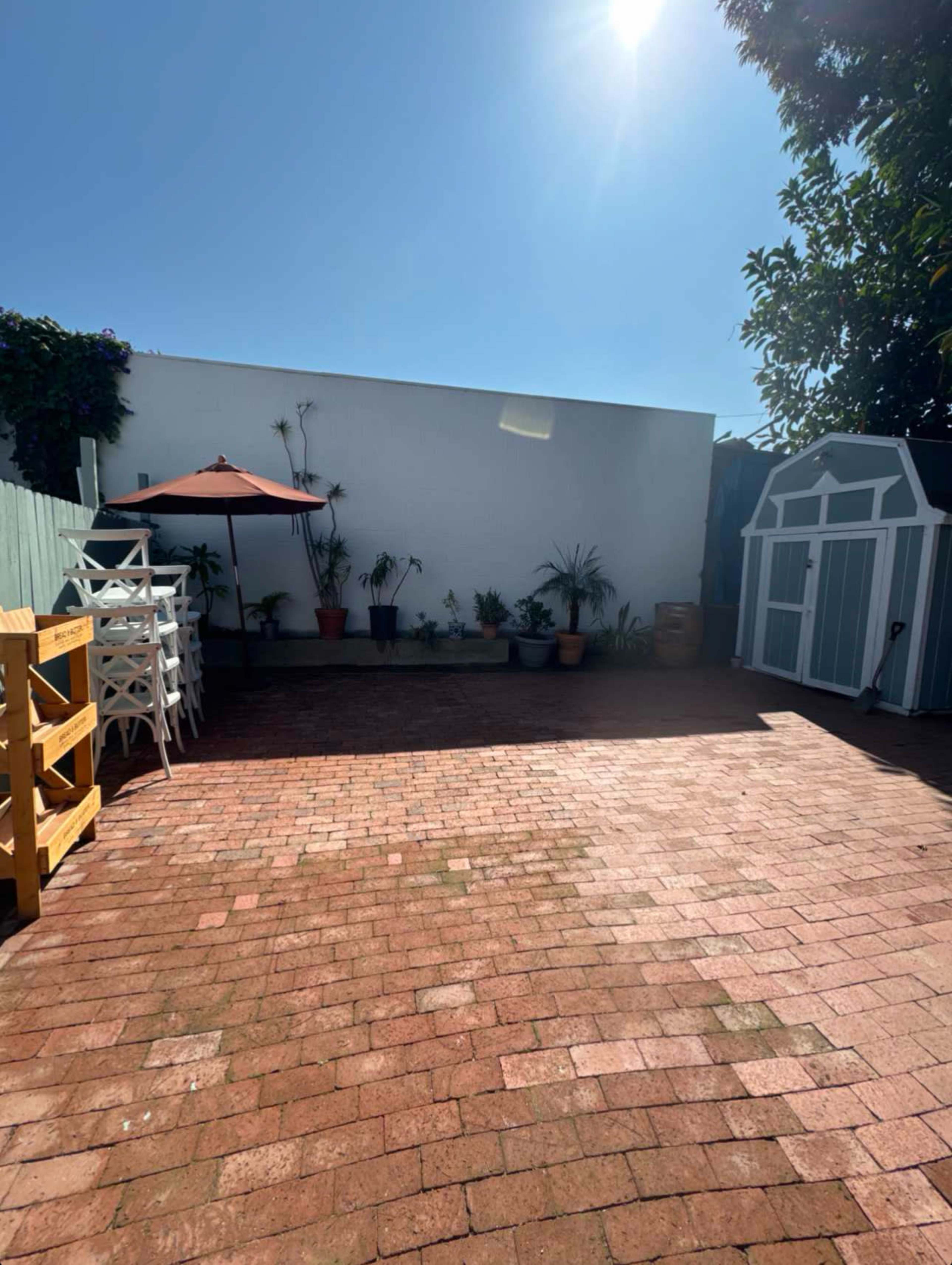The image shows a sunlit outdoor patio with brick flooring, an umbrella table, potted plants, and a small garden shed.