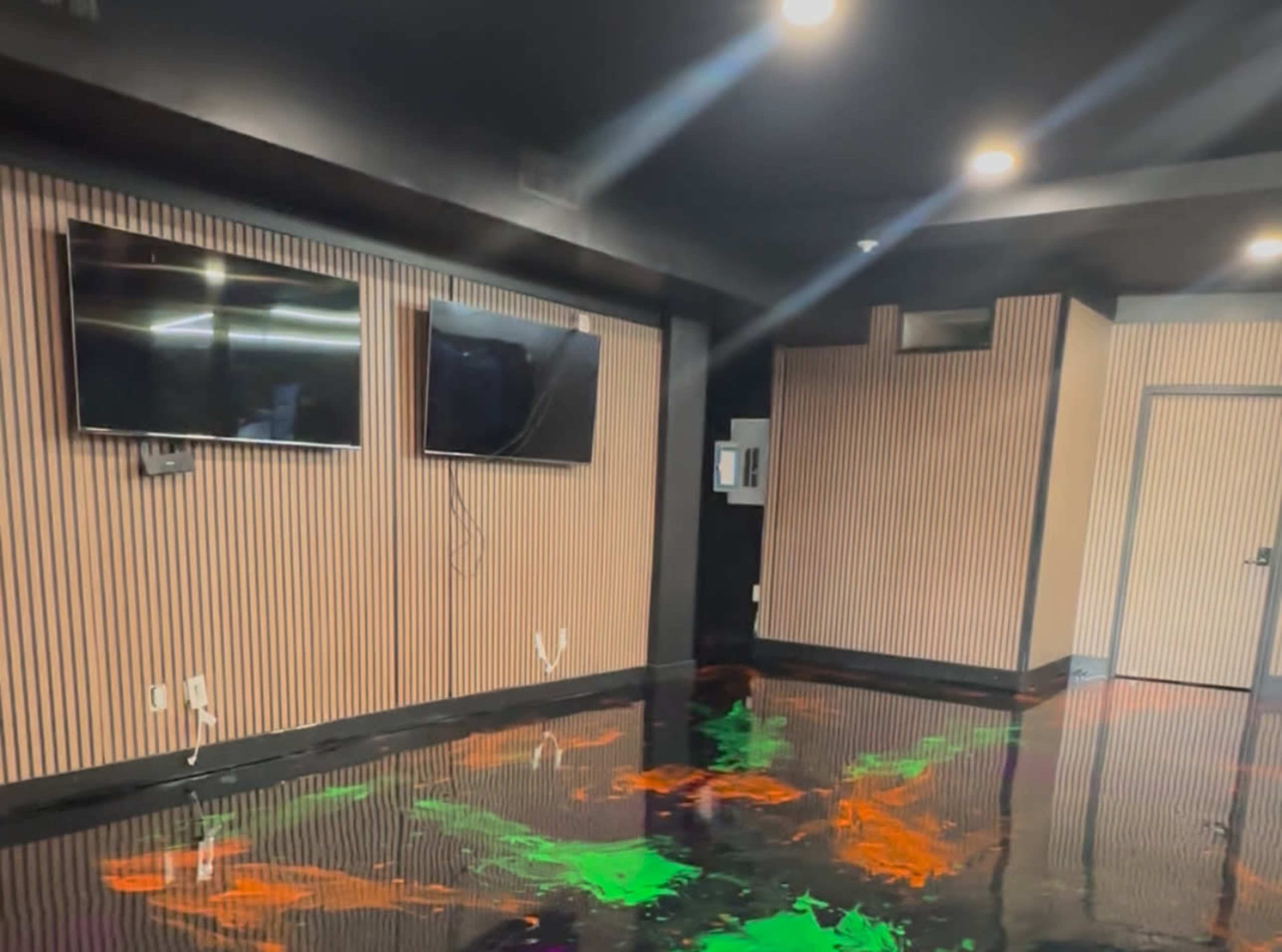 The image shows a modern room with dark walls, two wall-mounted televisions, and a glossy floor featuring colorful splashes of paint.