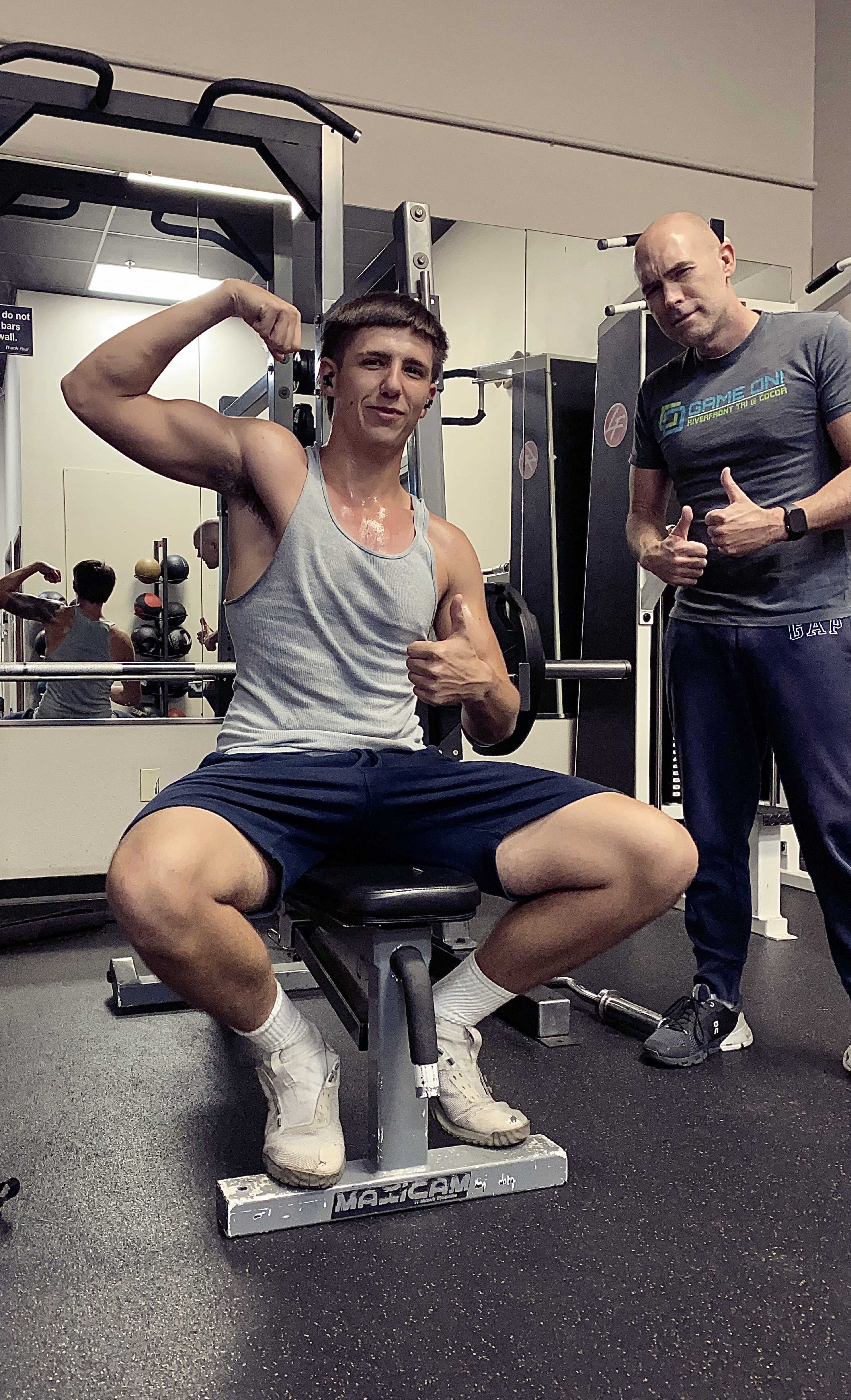 A young man in a sleeveless gray shirt poses with his arm flexed while seated on a weight bench in a gym, with a trainer giving a thumbs-up nearby.