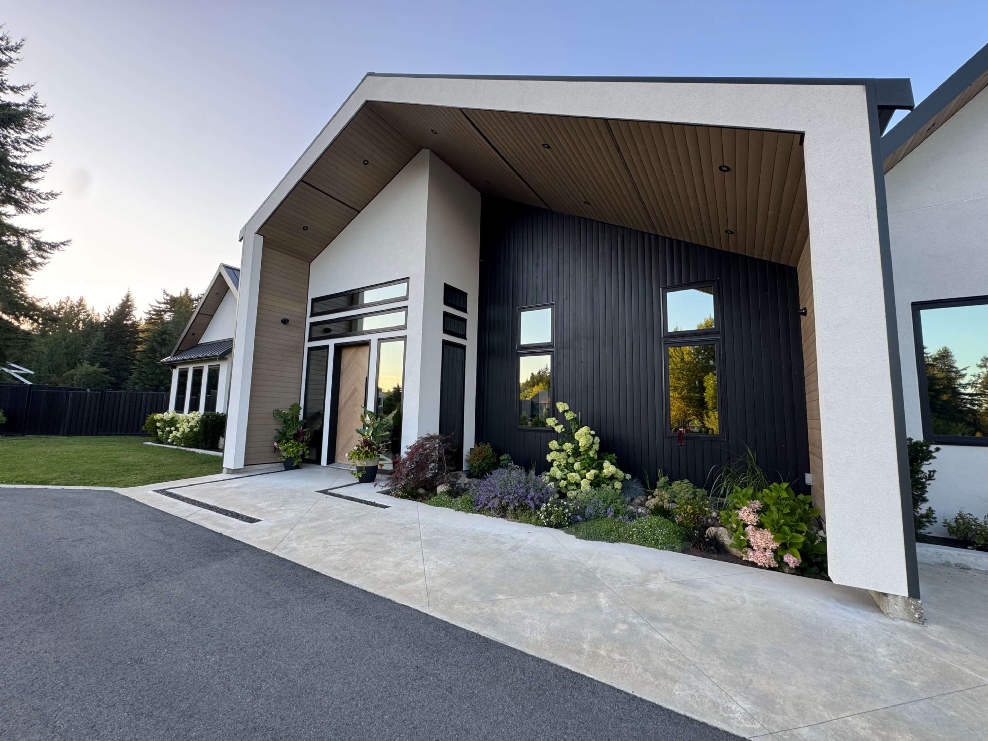 The image features a modern house with a distinctive angular roof, a mix of light and dark exterior walls, and a landscaped entrance with various plants and flowers.