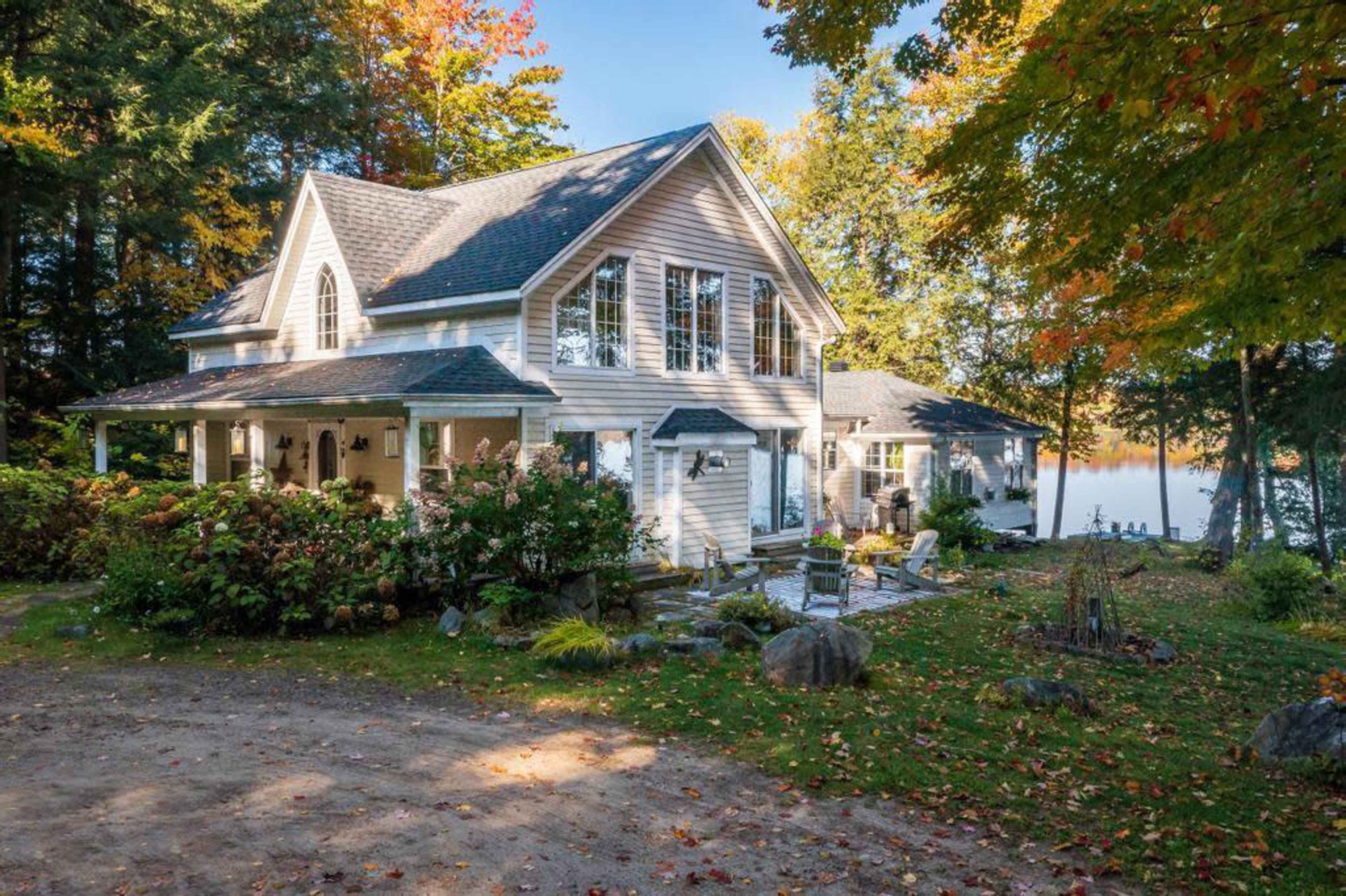 A two-story house with a wraparound porch is situated near a body of water, surrounded by trees with autumn foliage.