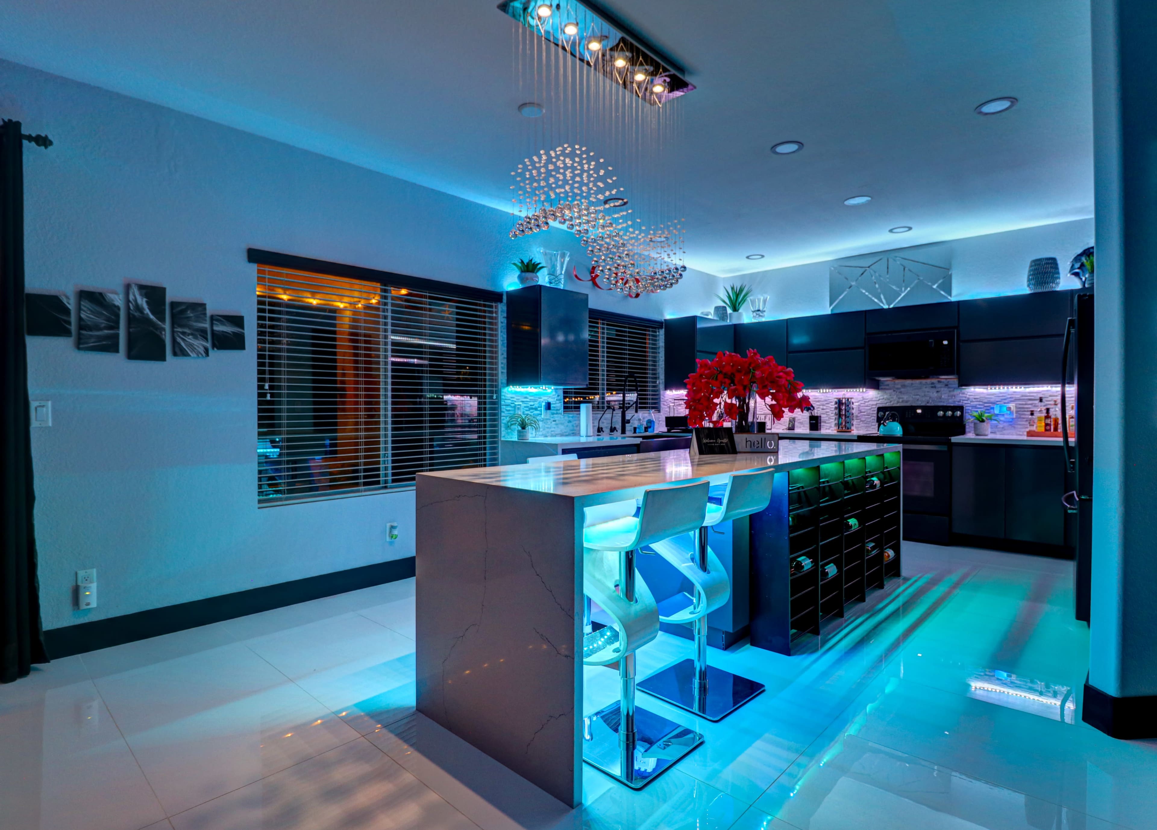 The image shows a modern kitchen with a central island, bar stools, and illuminated countertops, featuring a mix of blue and warm lighting.