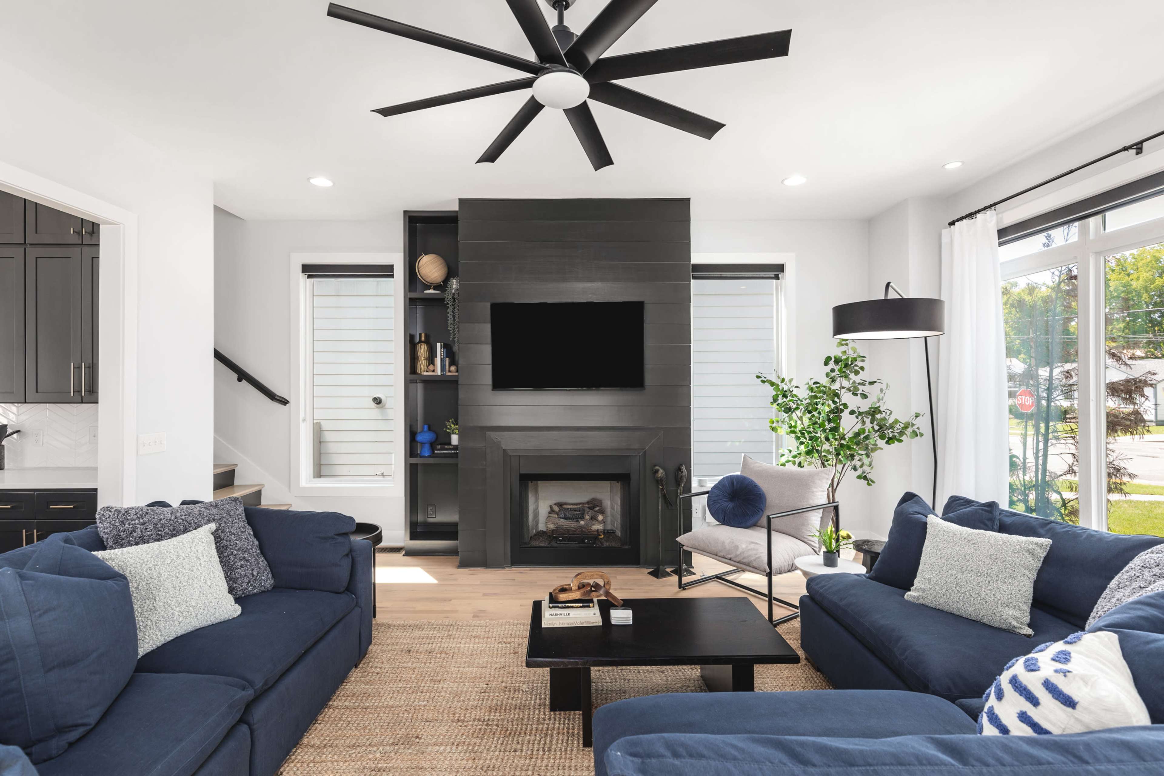 The living room features two blue sofas facing a black fireplace with a mounted television above it, surrounded by light walls and large windows.