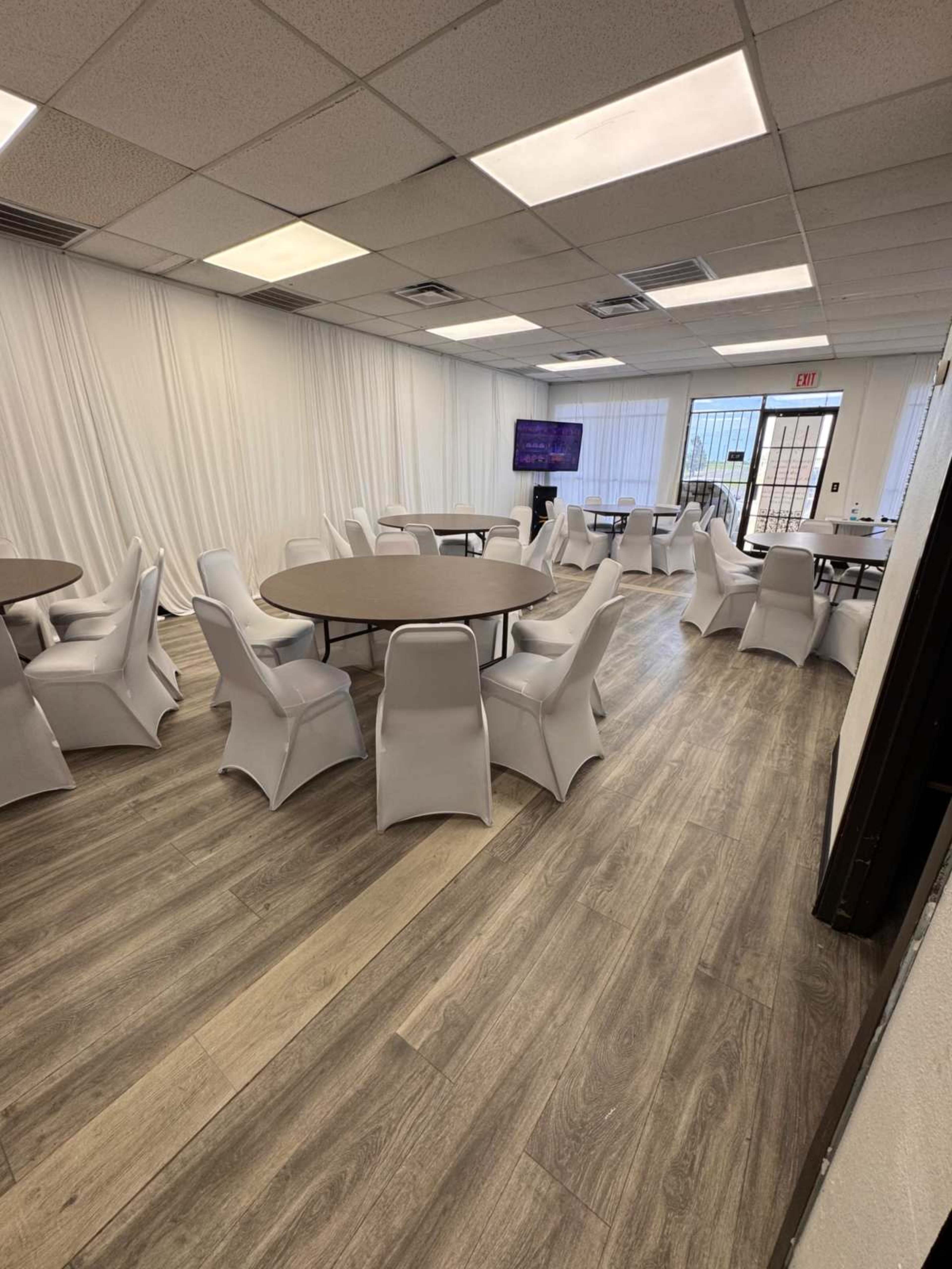 A spacious meeting room features circular tables surrounded by white chairs, with light-colored wood flooring and a large window letting in natural light.