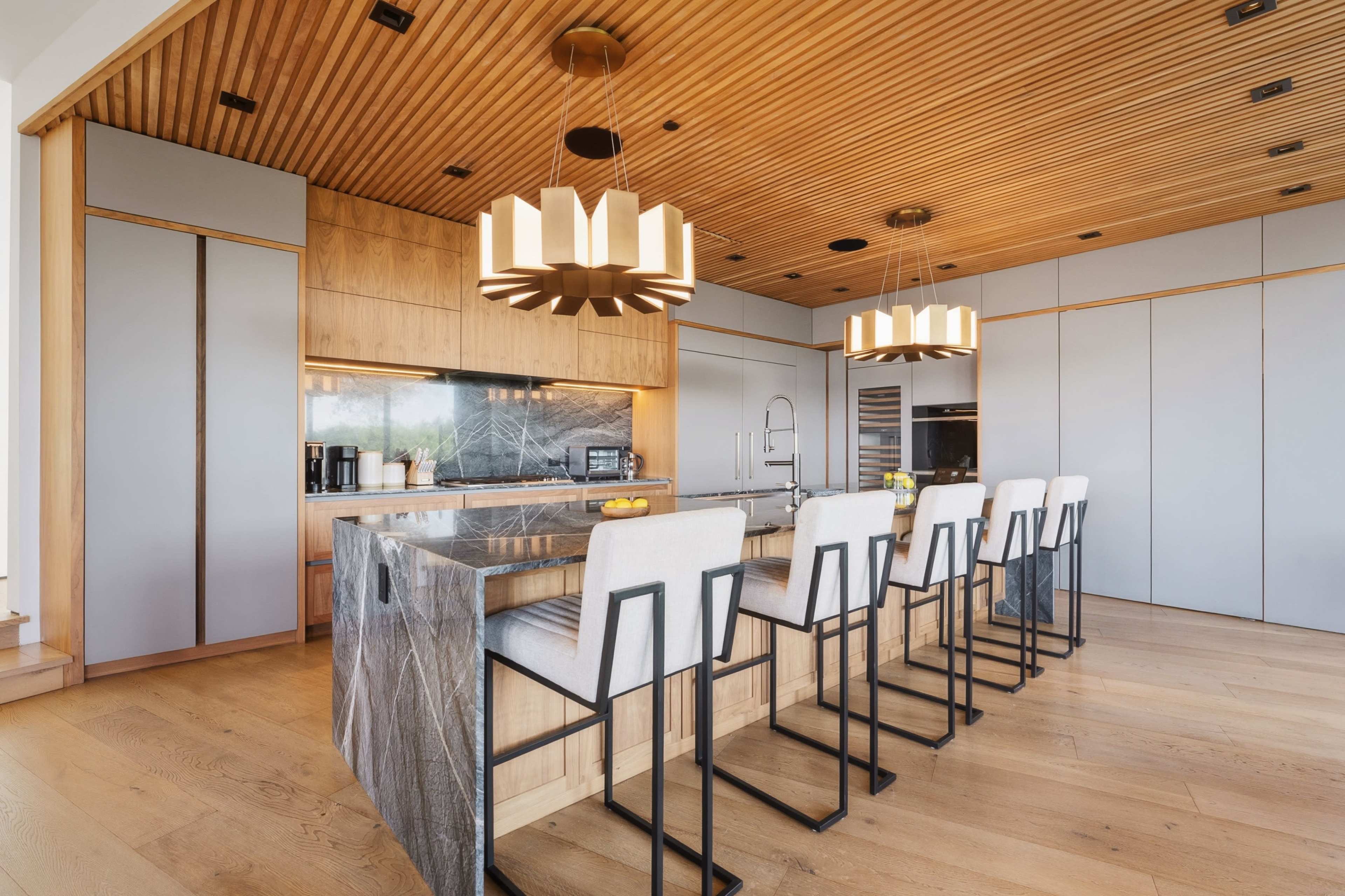 The image shows a modern kitchen with a large central island, high-backed bar stools, wooden ceiling beams, and stylish pendant lights.