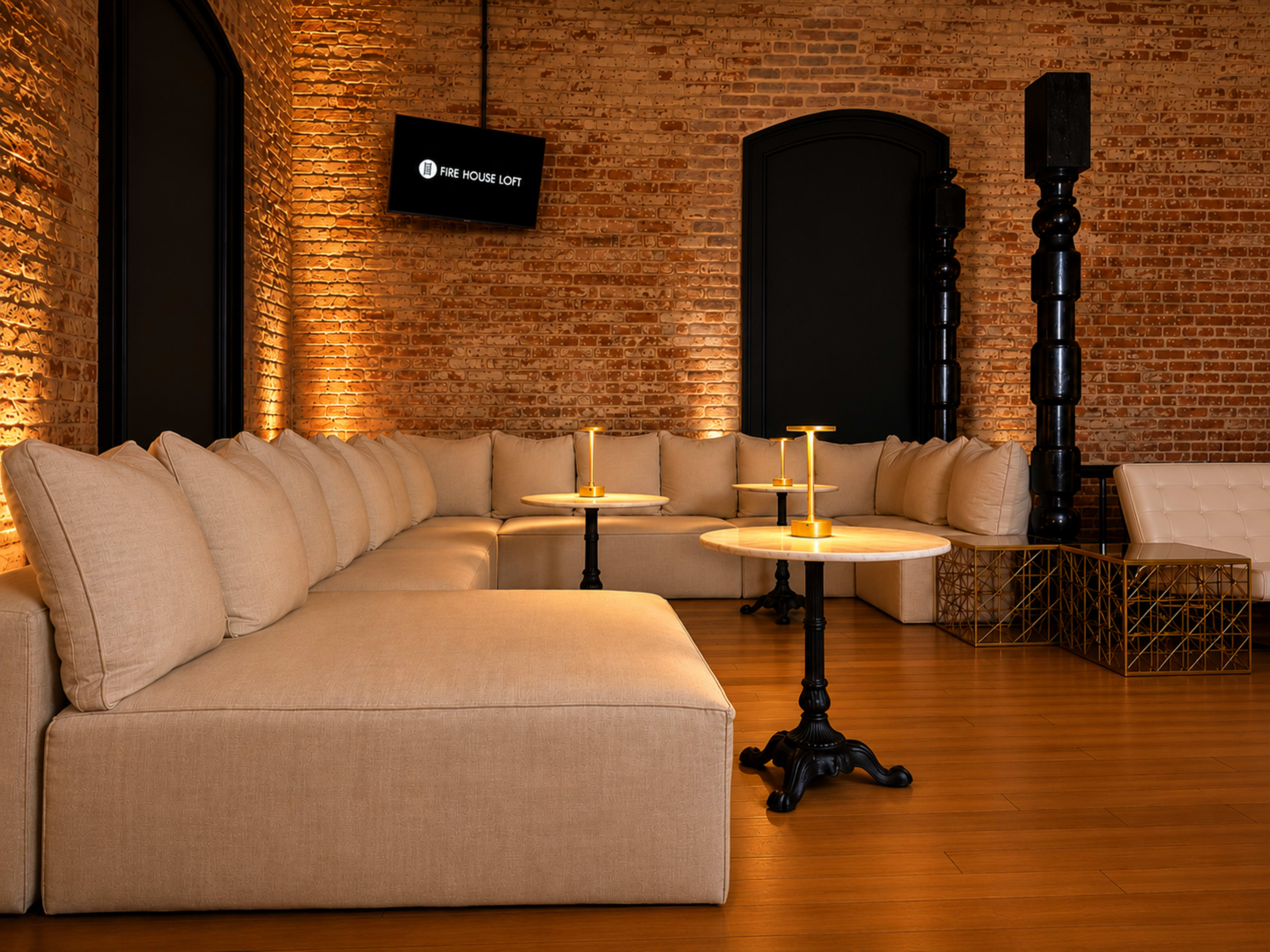 The image shows a stylish lounge area featuring a large, L-shaped beige couch, two circular tables with golden candle holders, and exposed brick walls illuminated by warm lighting.