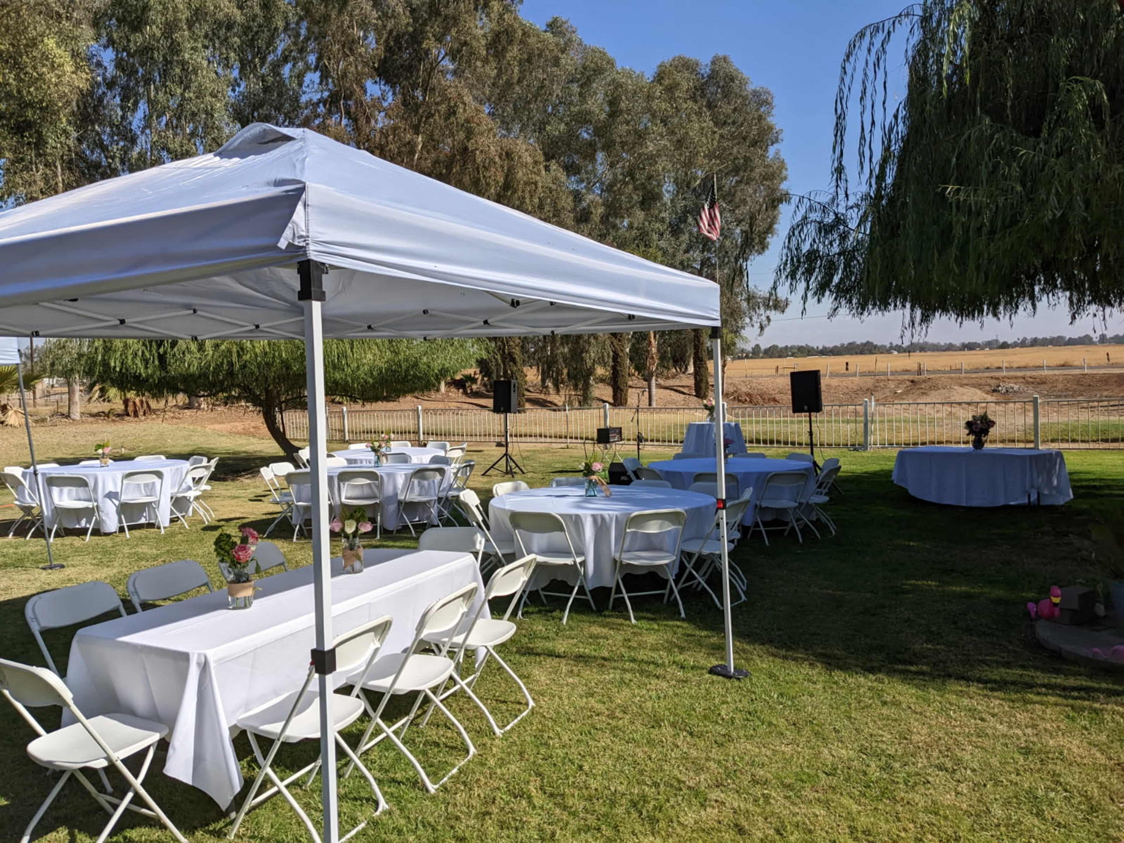 A white tent with tables and chairs is set up on a grassy area near a field, with trees and a flag in the background.