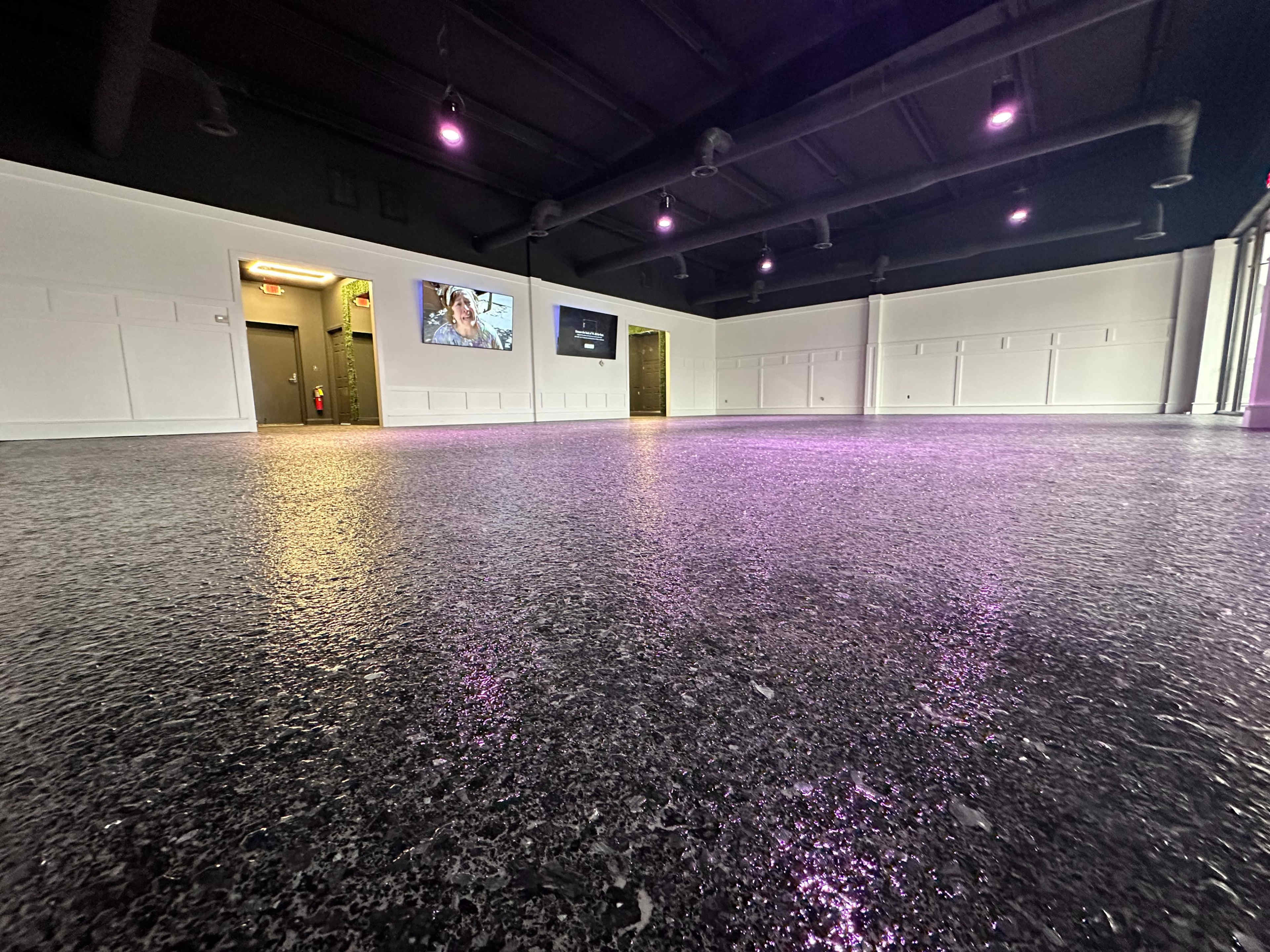 A spacious, brightly lit room with a polished floor, featuring purple-accented lighting and several doors leading to other areas.