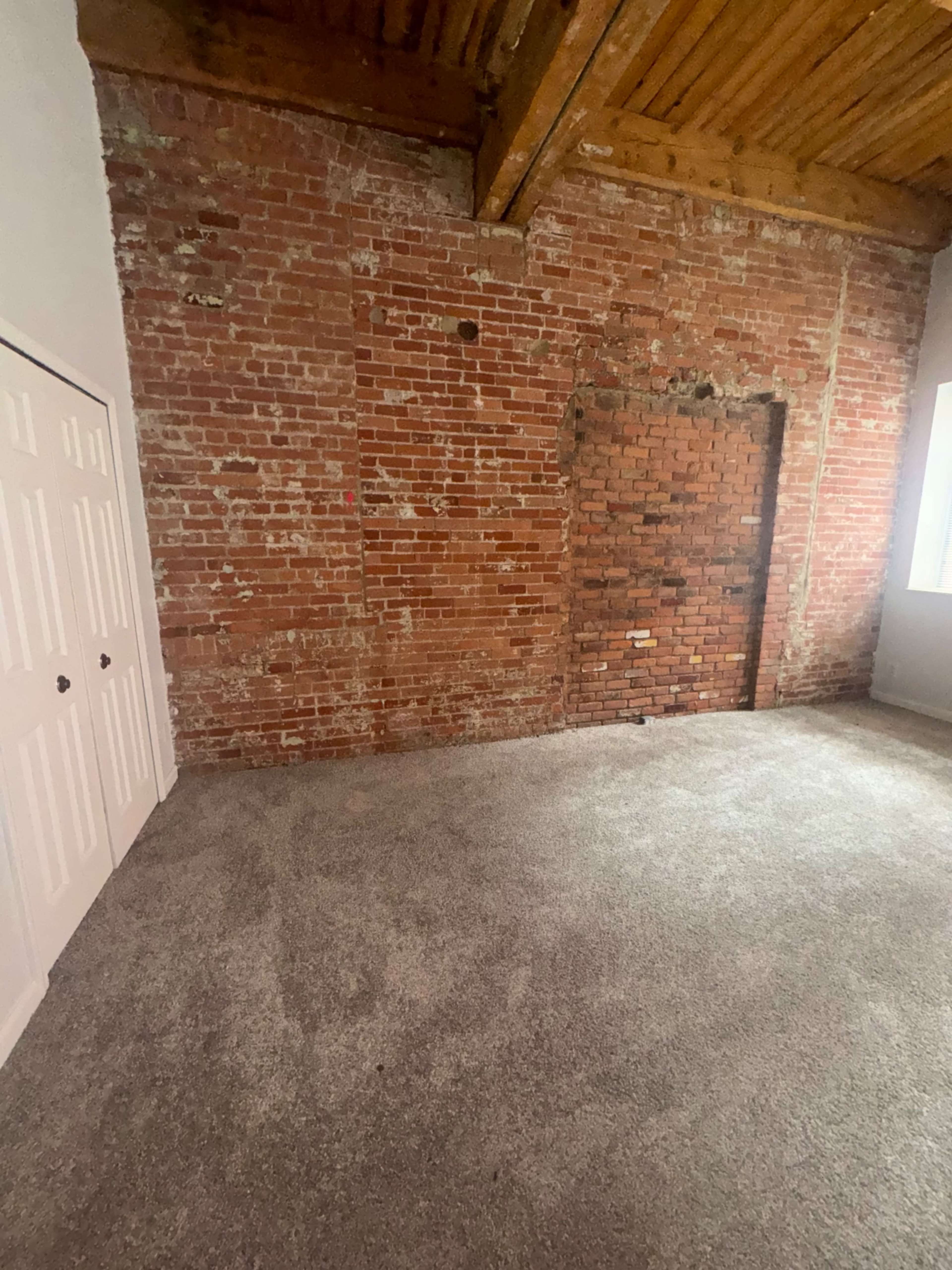 The image shows a room with carpeted flooring, exposed brick walls, and a partially bricked-up doorway.