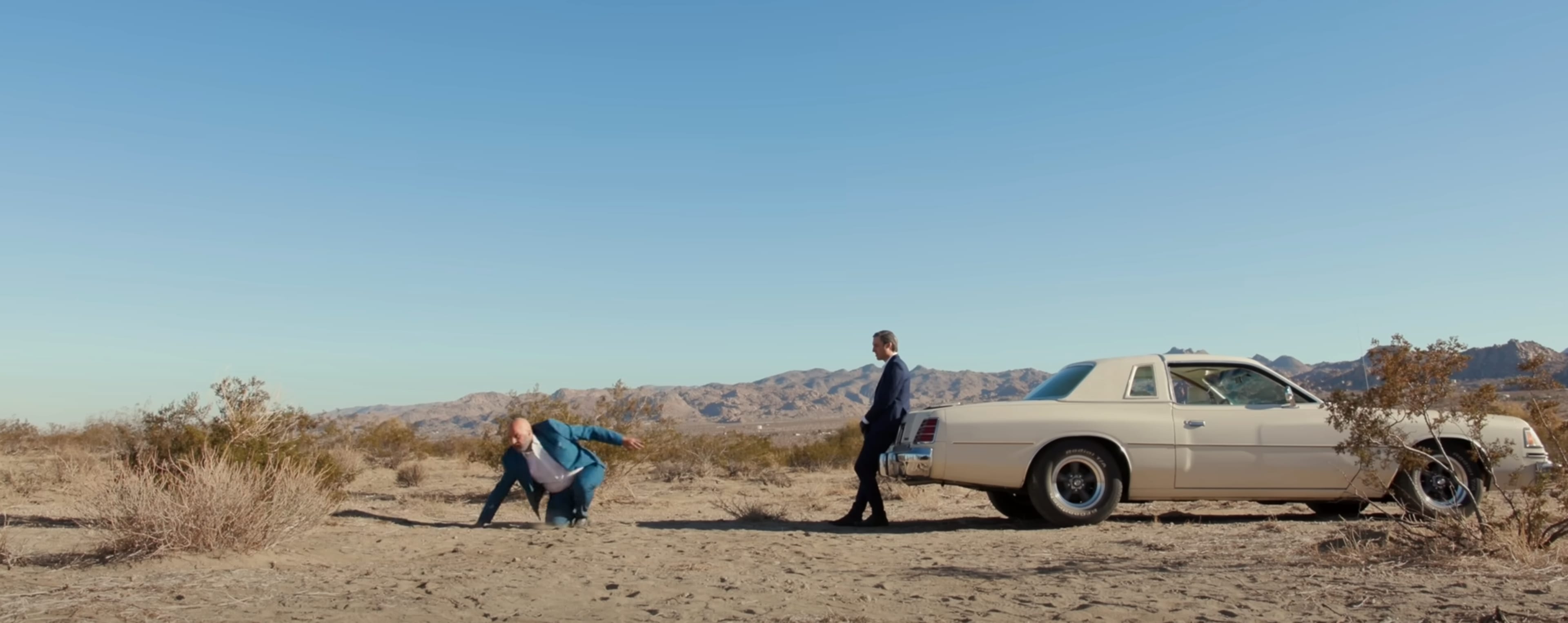 A man assists a woman who is kneeling on the ground near a vintage white car in a desert landscape with mountains in the background.
